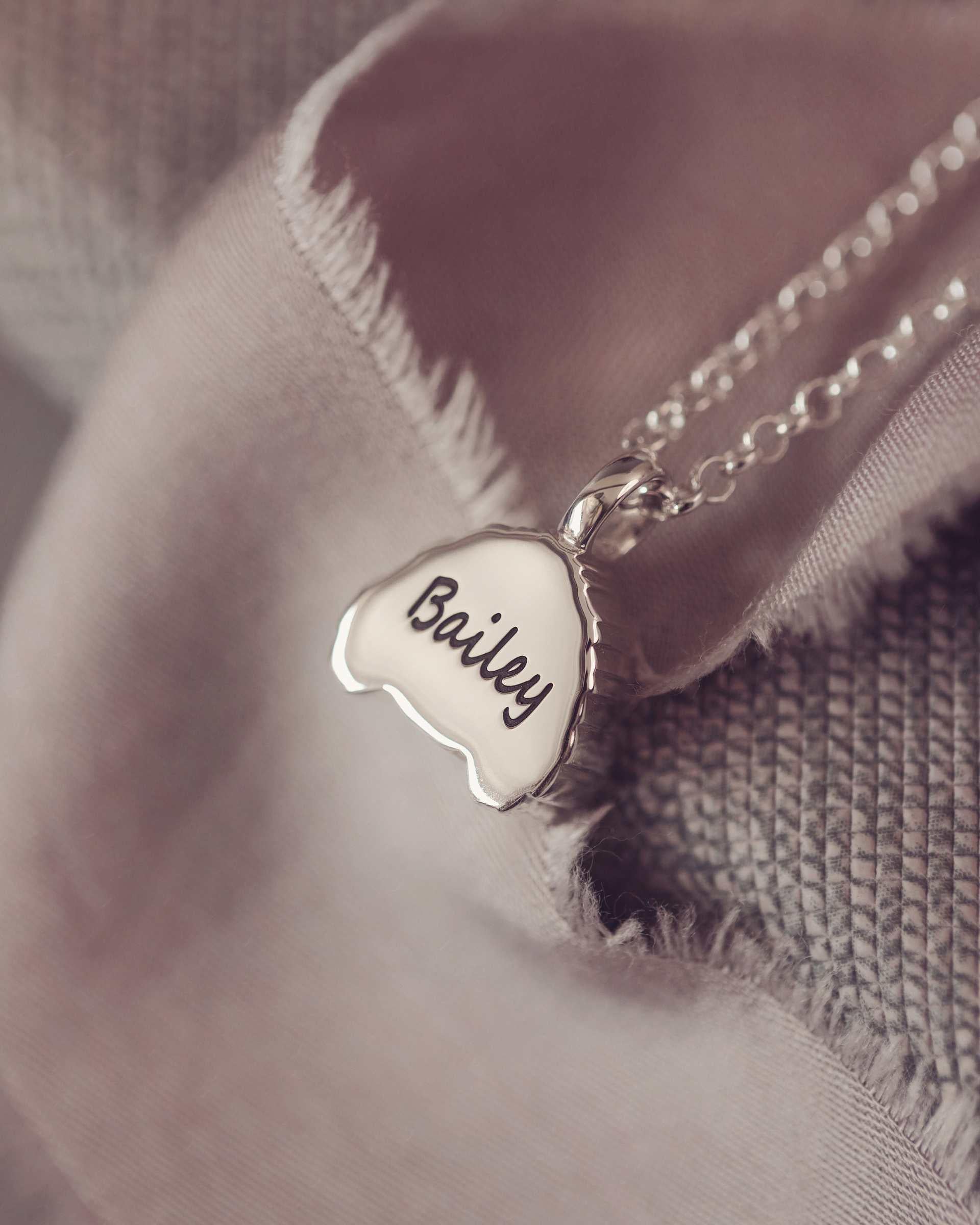 Silver necklace with a dog-shaped pendant engraved 'Bailey' on a soft fabric background