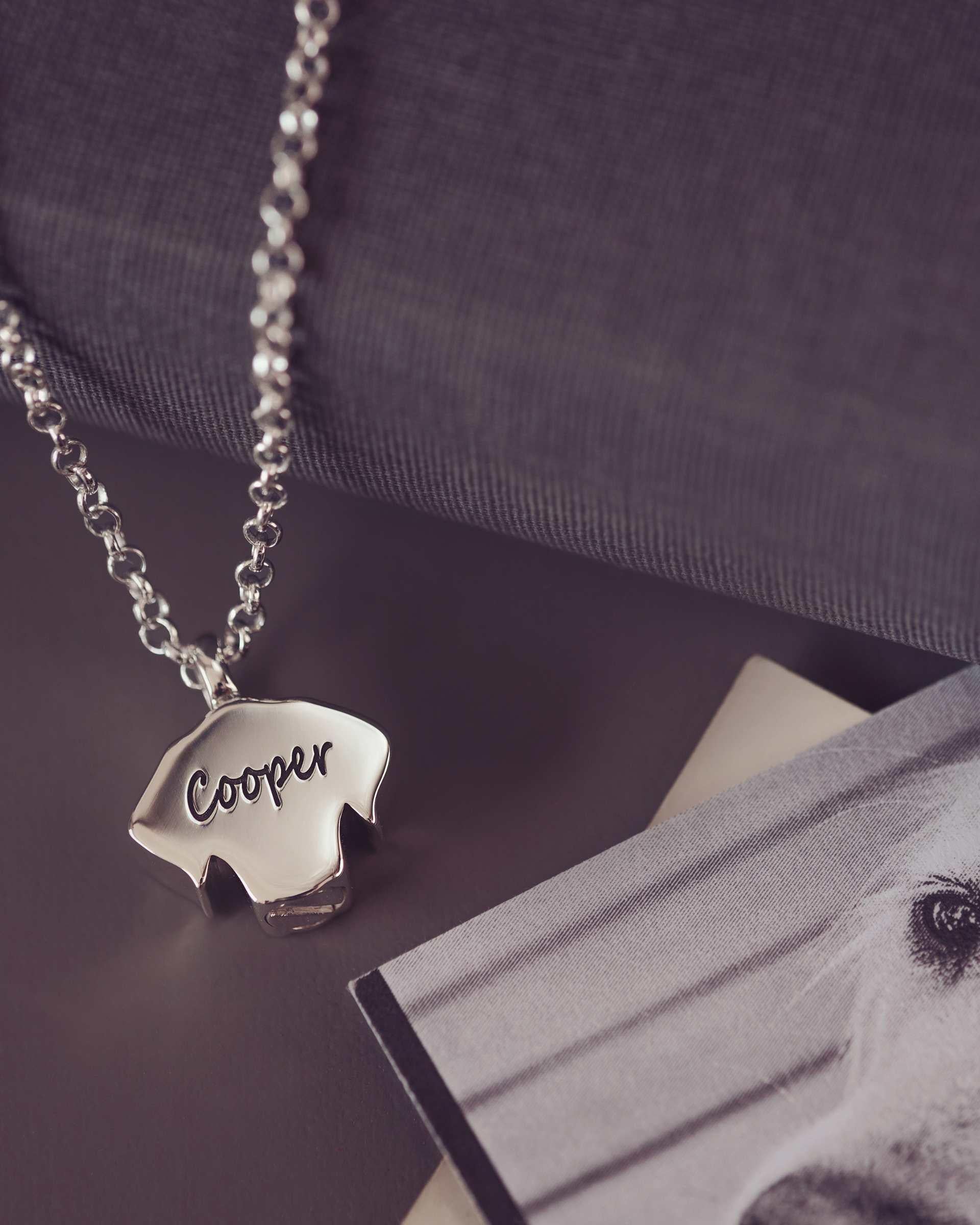 Silver necklace with a dog-shaped pendant displaying 'Cooper' on a dark surface with a photo of a dog.