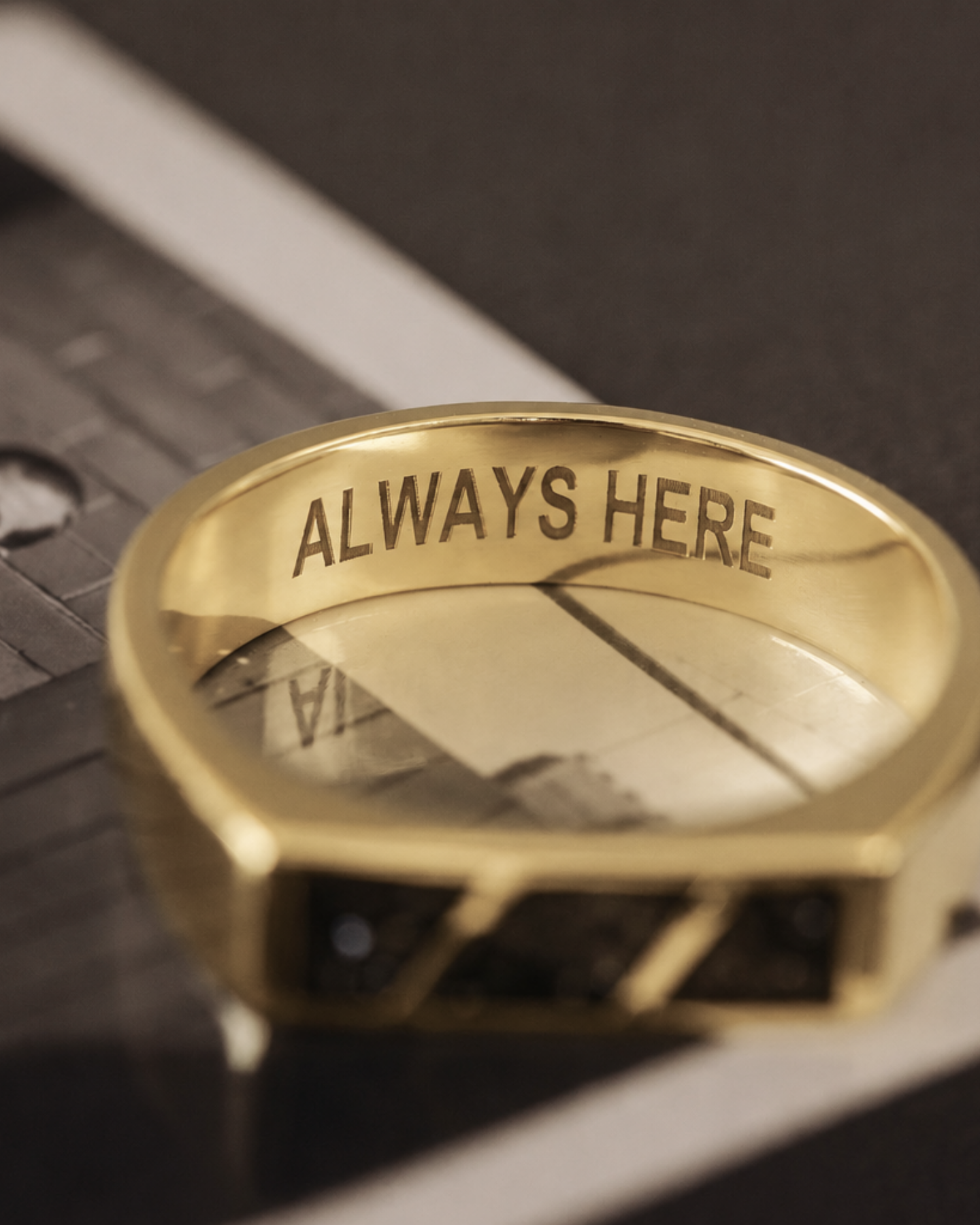 A gold-toned ring engraved with the words 'ALWAYS HERE' on the inside band, resting on a black-and-white printed card over a dark surface.