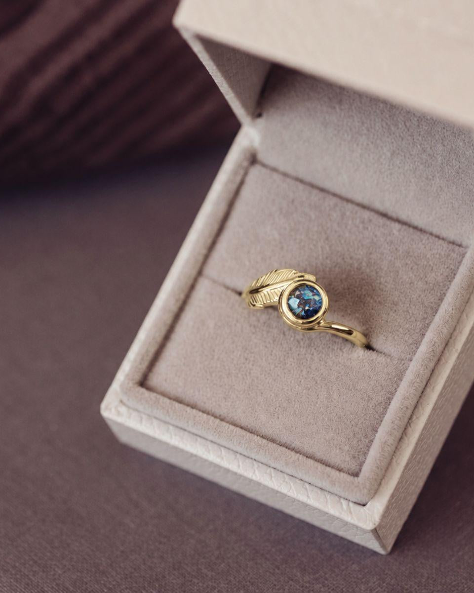 A gold ring with a textured leaf-shaped setting and a round blue gemstone, displayed inside an open beige jewelry box on a dark brown surface.