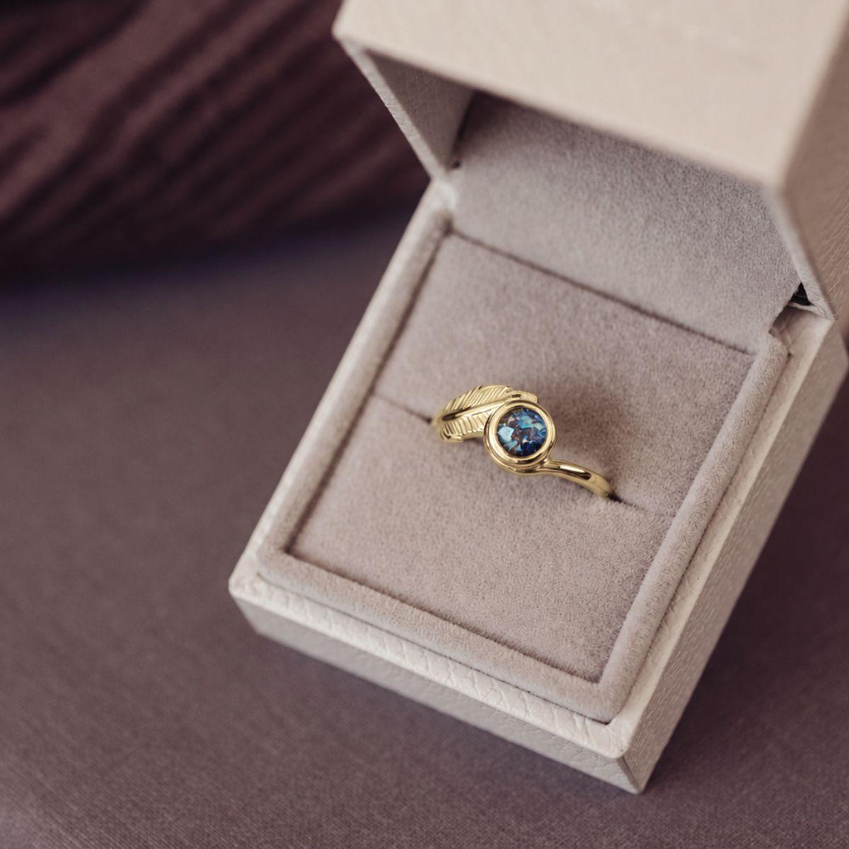 A gold ring with a textured leaf-shaped setting and a round blue gemstone, displayed inside an open beige jewelry box on a dark brown surface.