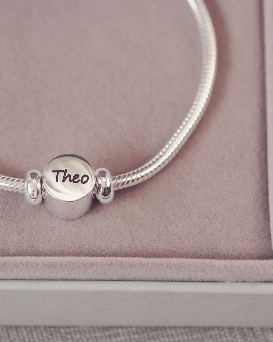 Silver bracelet with a round charm engraved with 'Theo' on a pink cushioned surface.