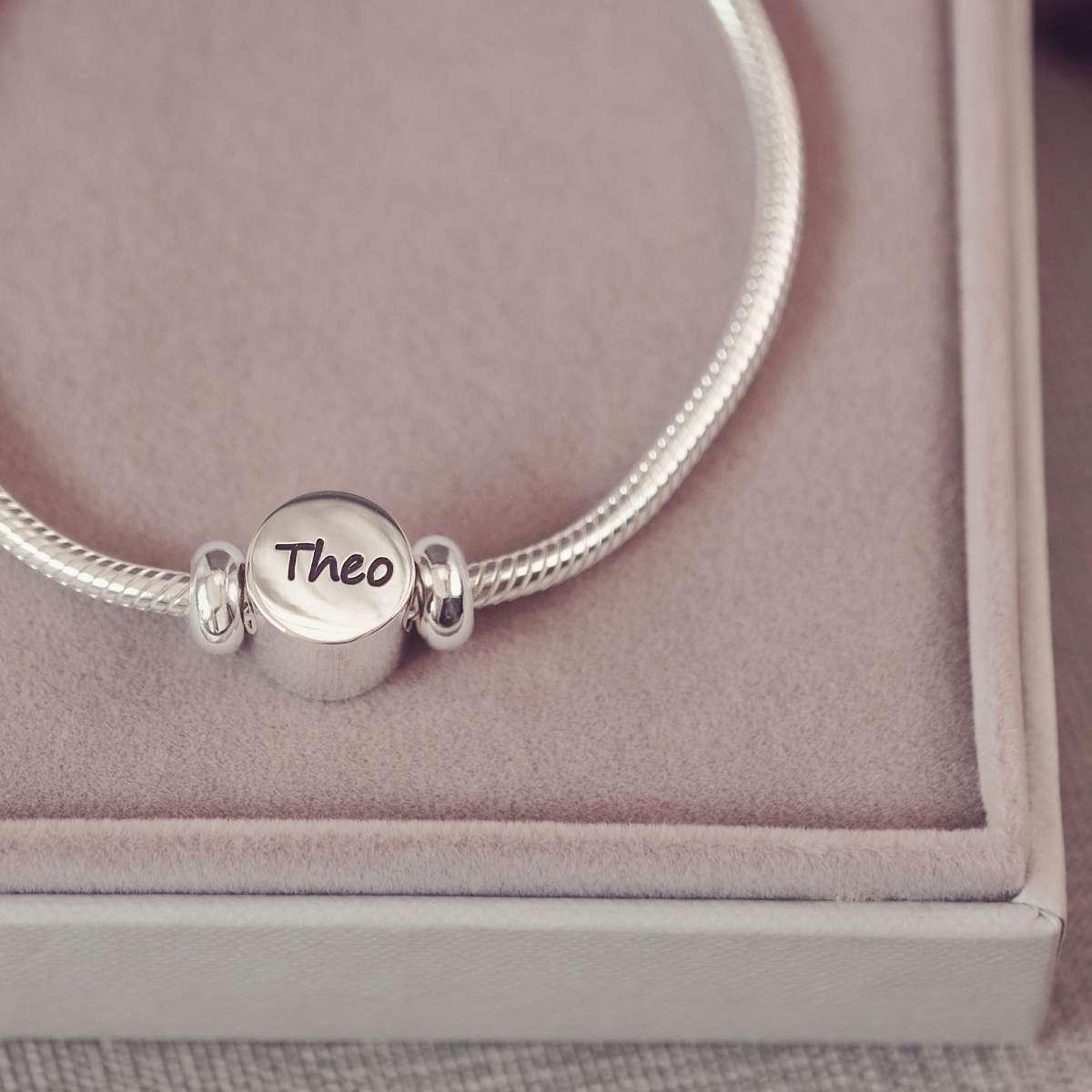 Silver bracelet with a round charm engraved with 'Theo' on a pink cushioned surface.