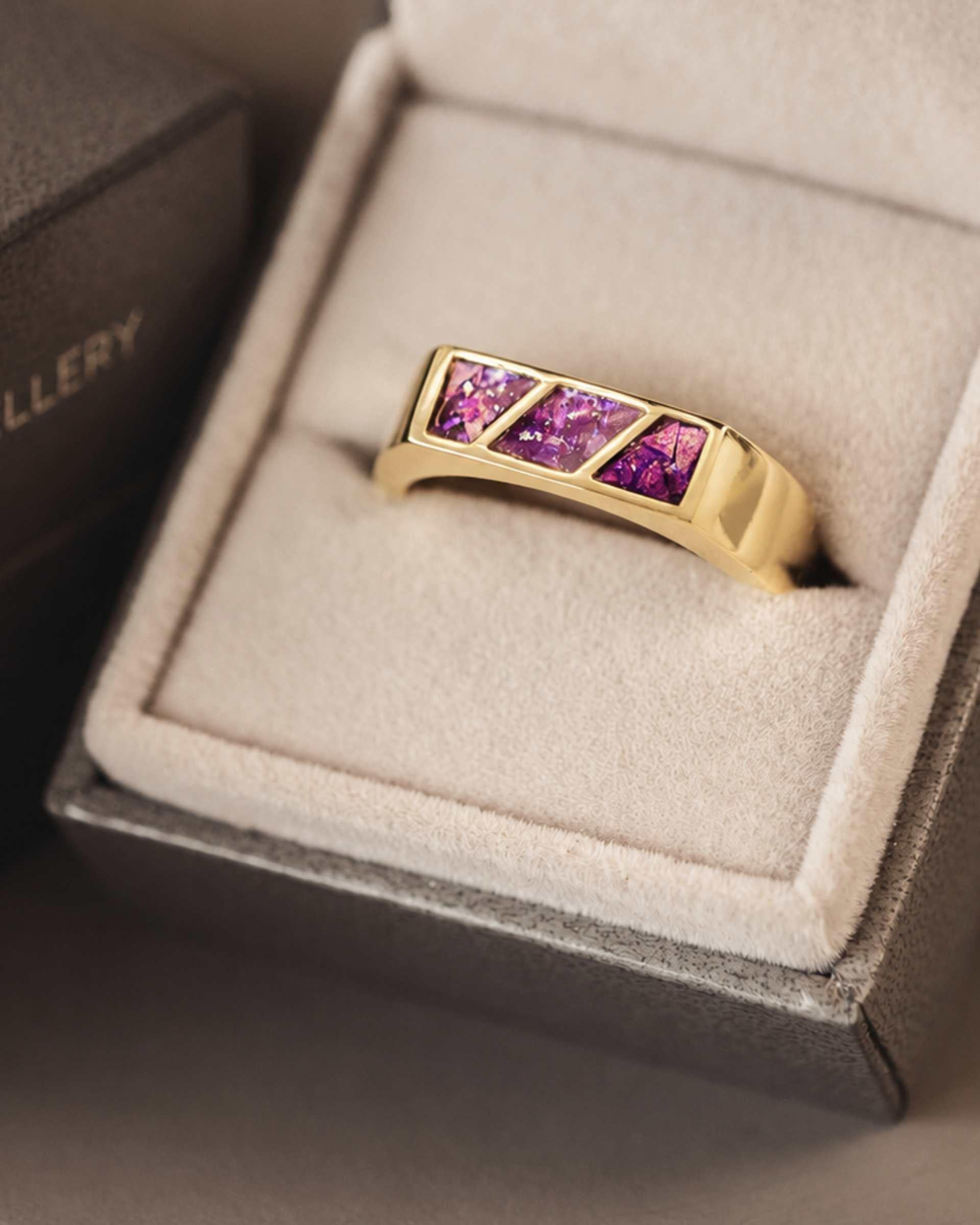 A gold ring with 3 sections of purple resin across the top sitting in an open jewellery box.