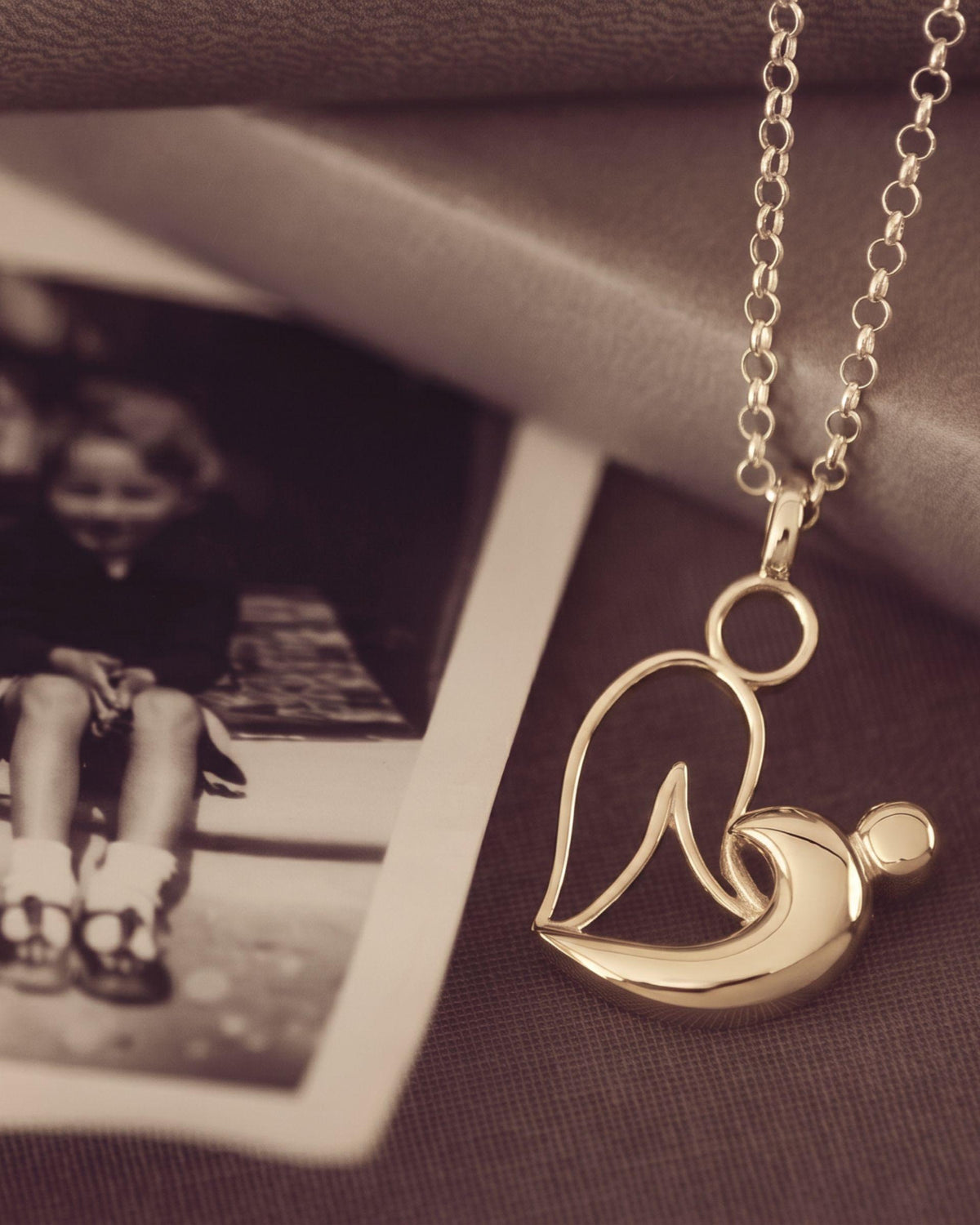 A gold pendant necklace featuring a stylized mother-and-child silhouette design, displayed on a dark textured surface next to a vintage sepia-toned photograph.