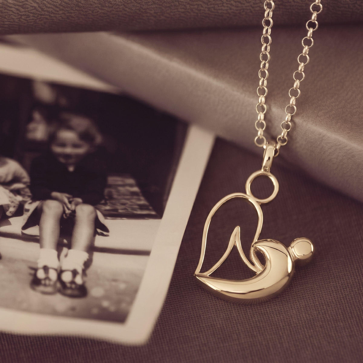 A gold pendant necklace featuring a stylized mother-and-child silhouette design, displayed on a dark textured surface next to a vintage sepia-toned photograph.