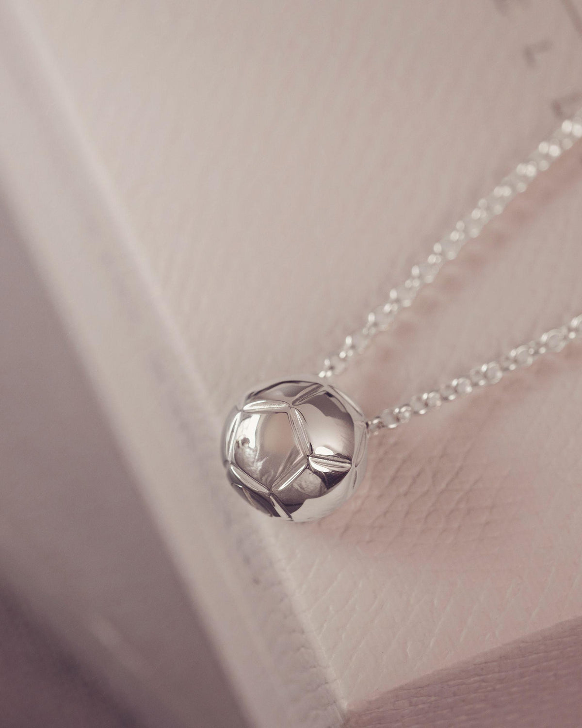 A close-up of a silver pendant necklace with a football, geometric ball design on a delicate chain, resting on a textured light pink surface.