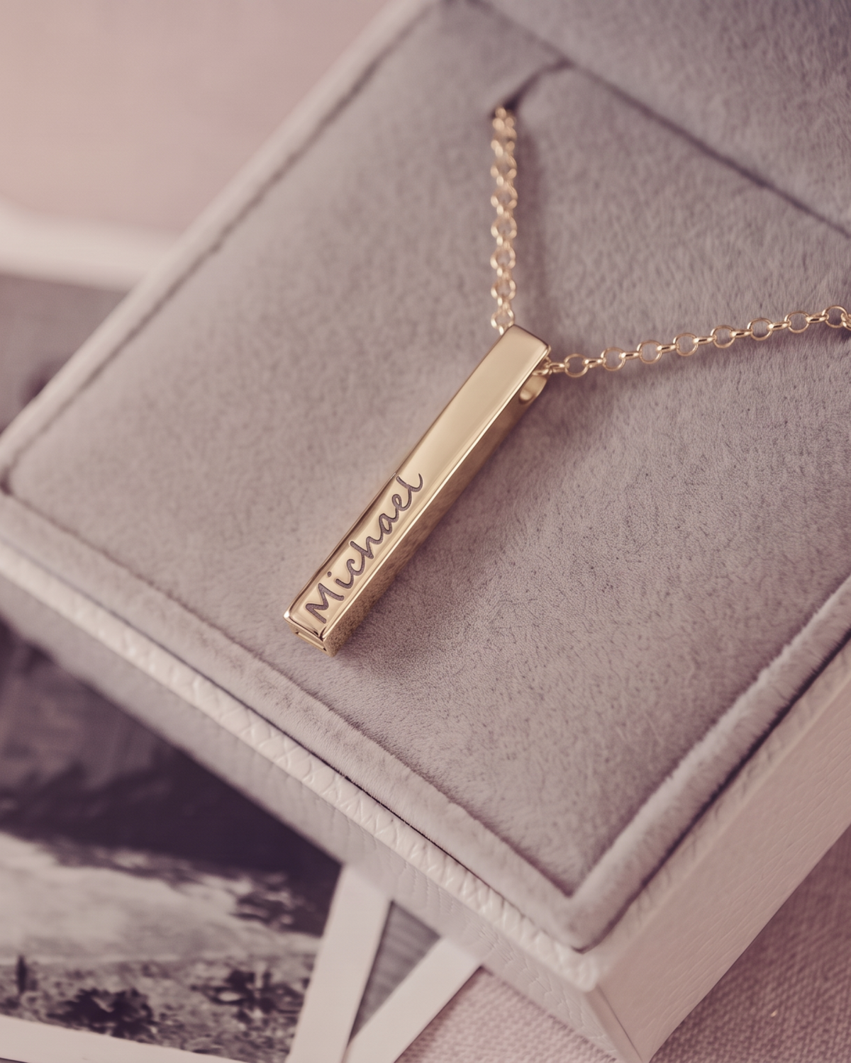 A gold bar necklace engraved with the name 'Michael' in script, resting on a gray velvet jewelry box.