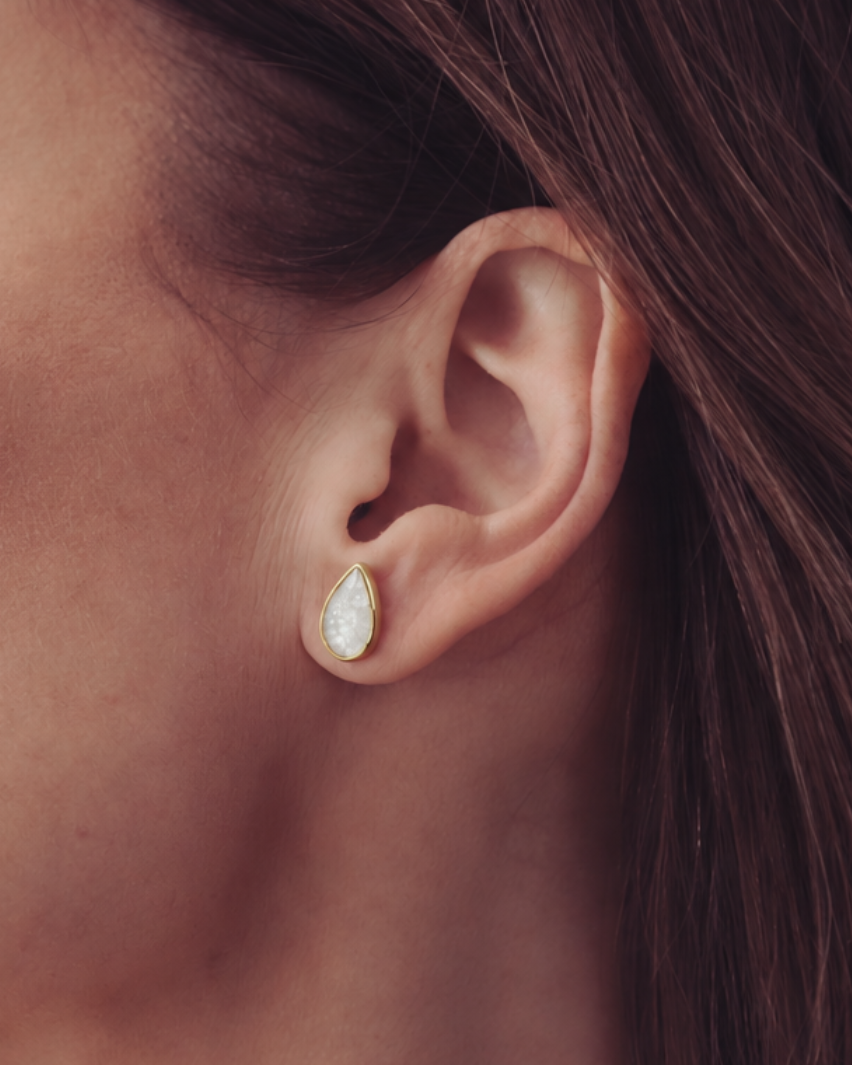 A close-up of a woman's ear wearing a small teardrop-shaped stud earring with a white iridescent center and a gold-toned bezel.