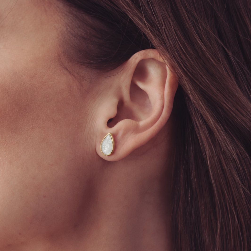 A close-up of a woman's ear wearing a small teardrop-shaped stud earring with a white iridescent center and a gold-toned bezel.