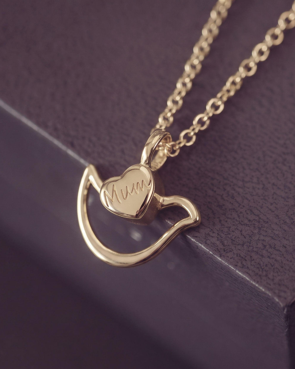 A gold necklace with a Dove & heart-shaped pendant engraved with 'Mum', resting on a dark textured surface.