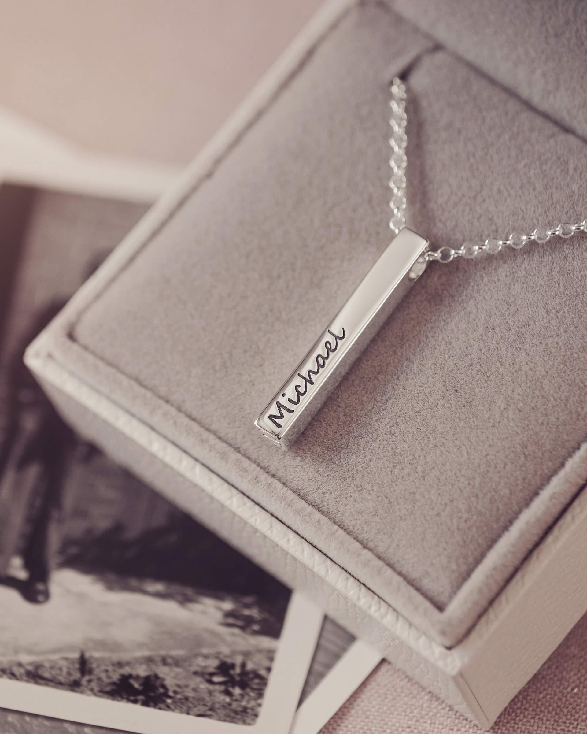 Silver necklace with 'Michael' engraving on a textured surface with blurred black and white photos in the background.