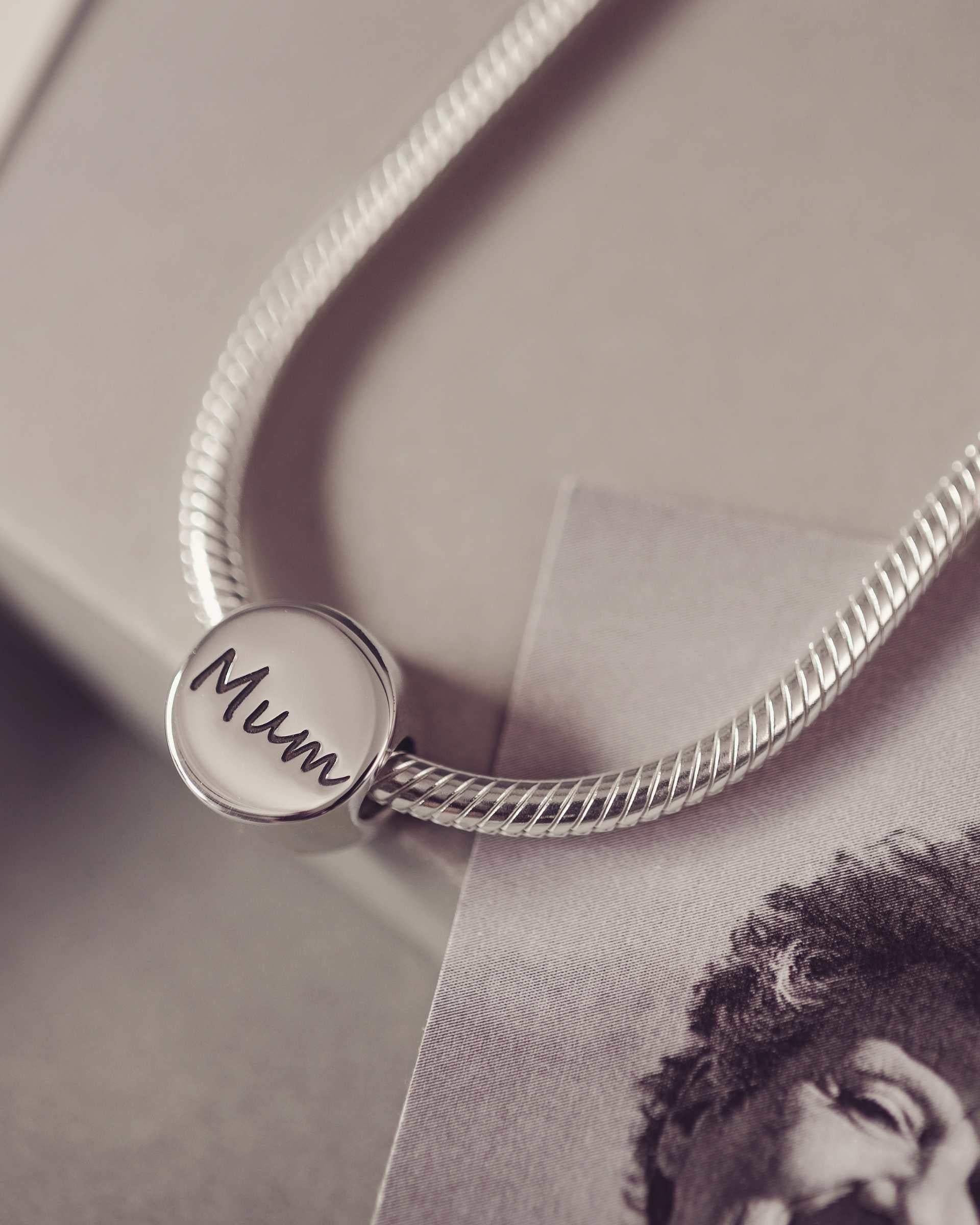 Round bracelet charm engraved with 'Mum' sitting on a thick snake chain. Grey background along side a black and white photograph.