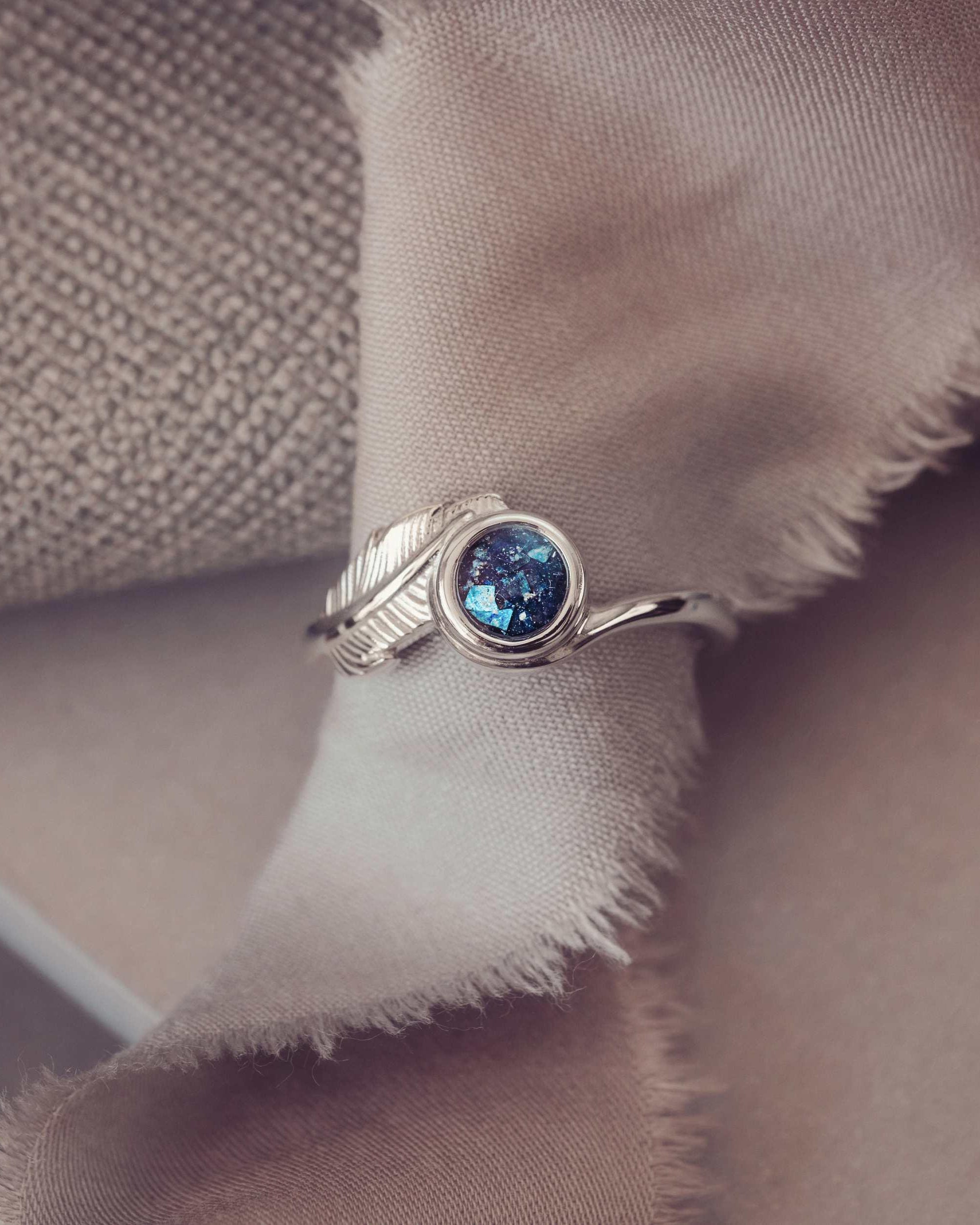 A silver ash filled feather ring sitting around a piece of cloth on a neutral backgound.