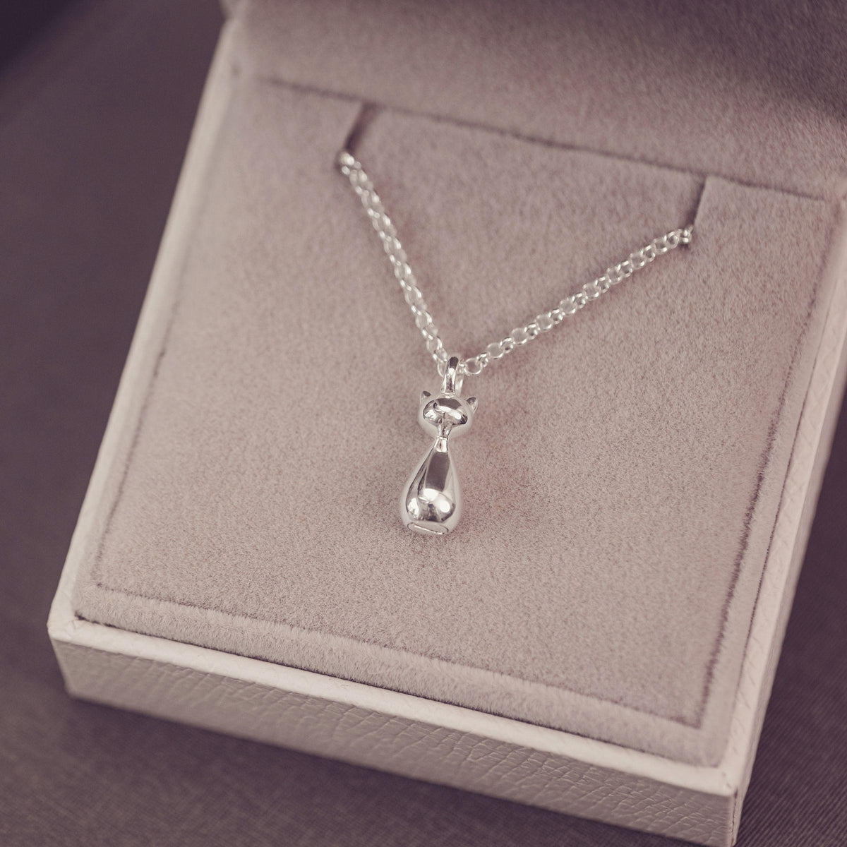 A stylised silver cat pendant necklace on a delicate chain, displayed inside a light beige jewelry box.