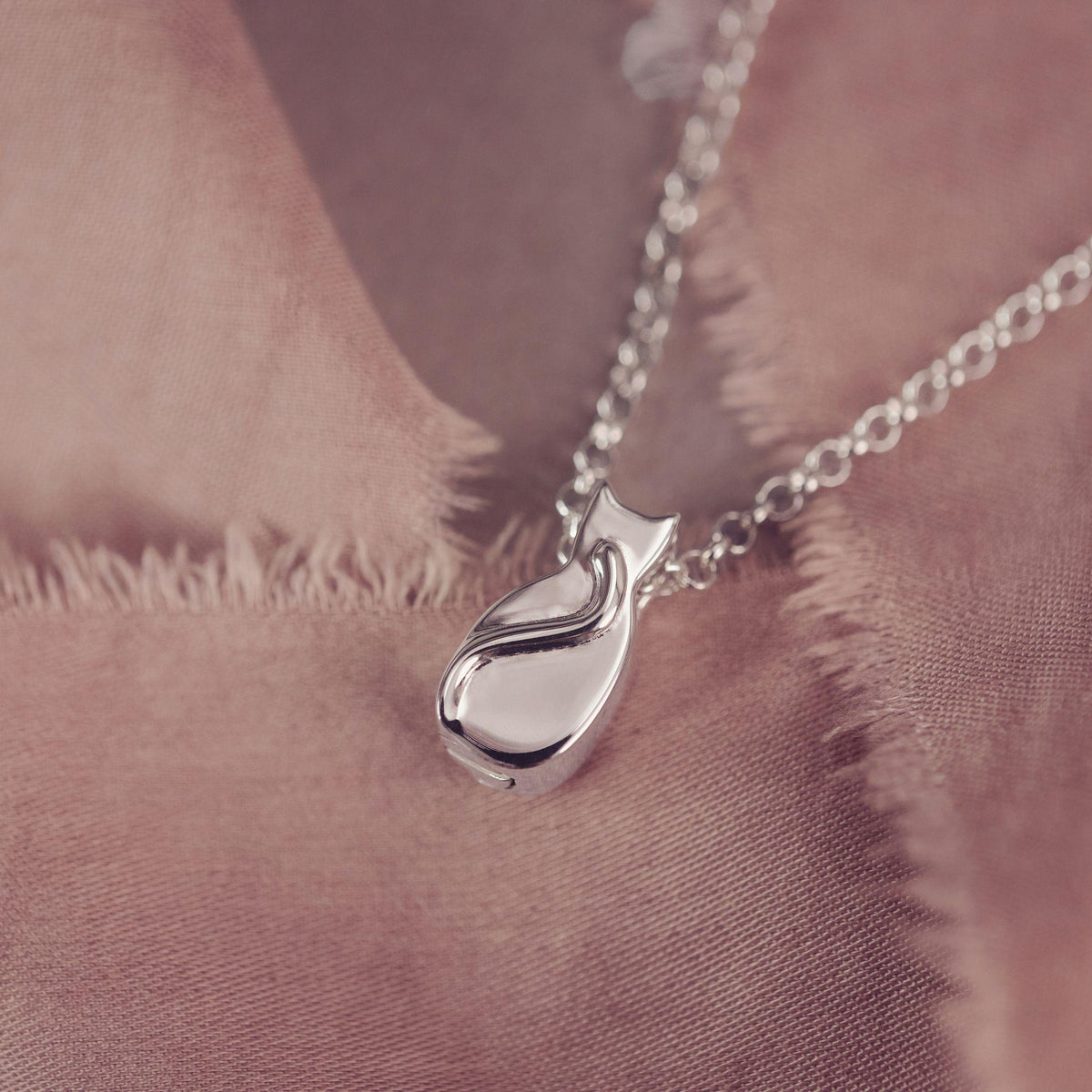 A polished silver pendant necklace with a Cat style pendant a soft, dusty rose textured fabric.