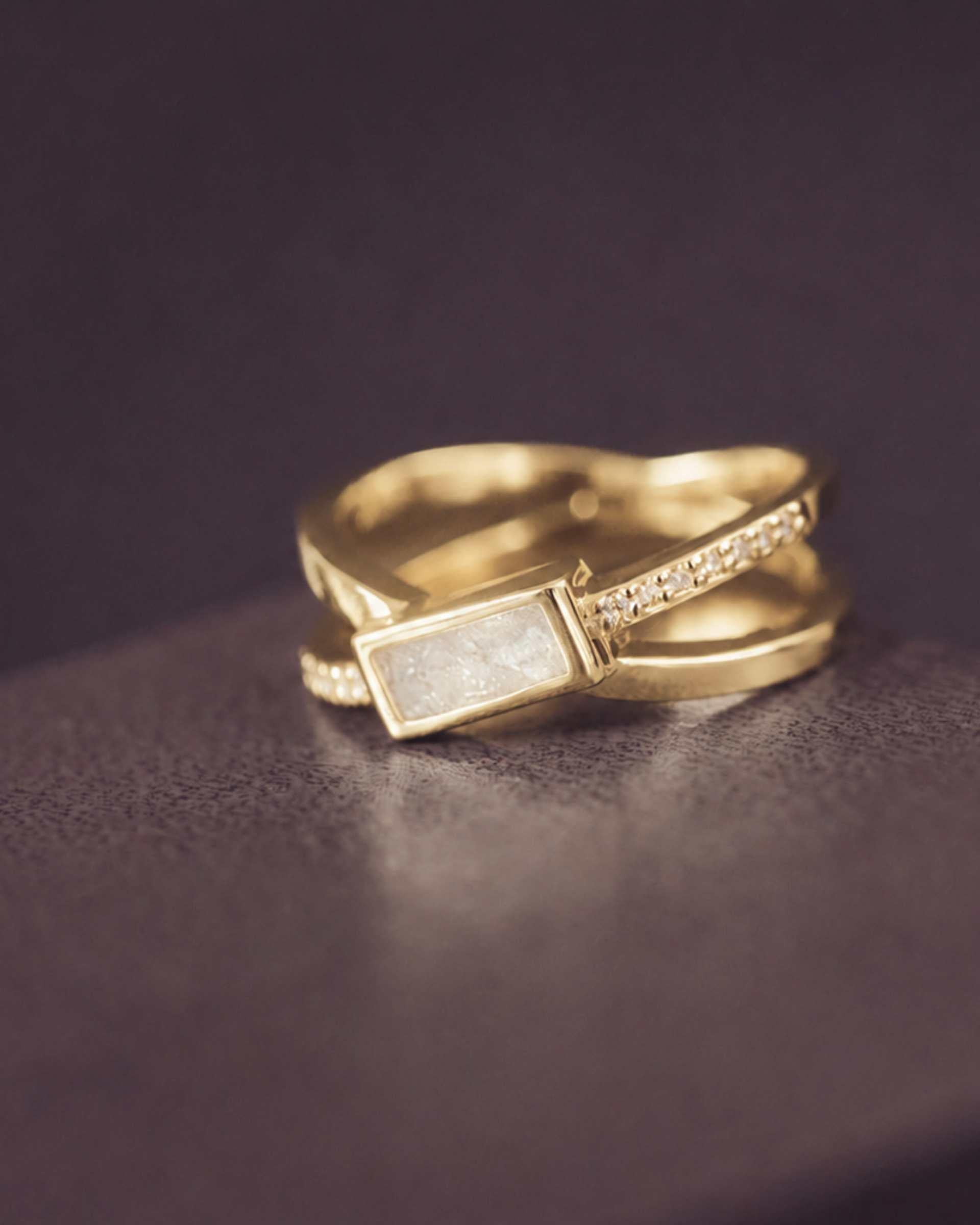 gold criss cross ring with a white rectangle ash stone and gemstones on a dark background
