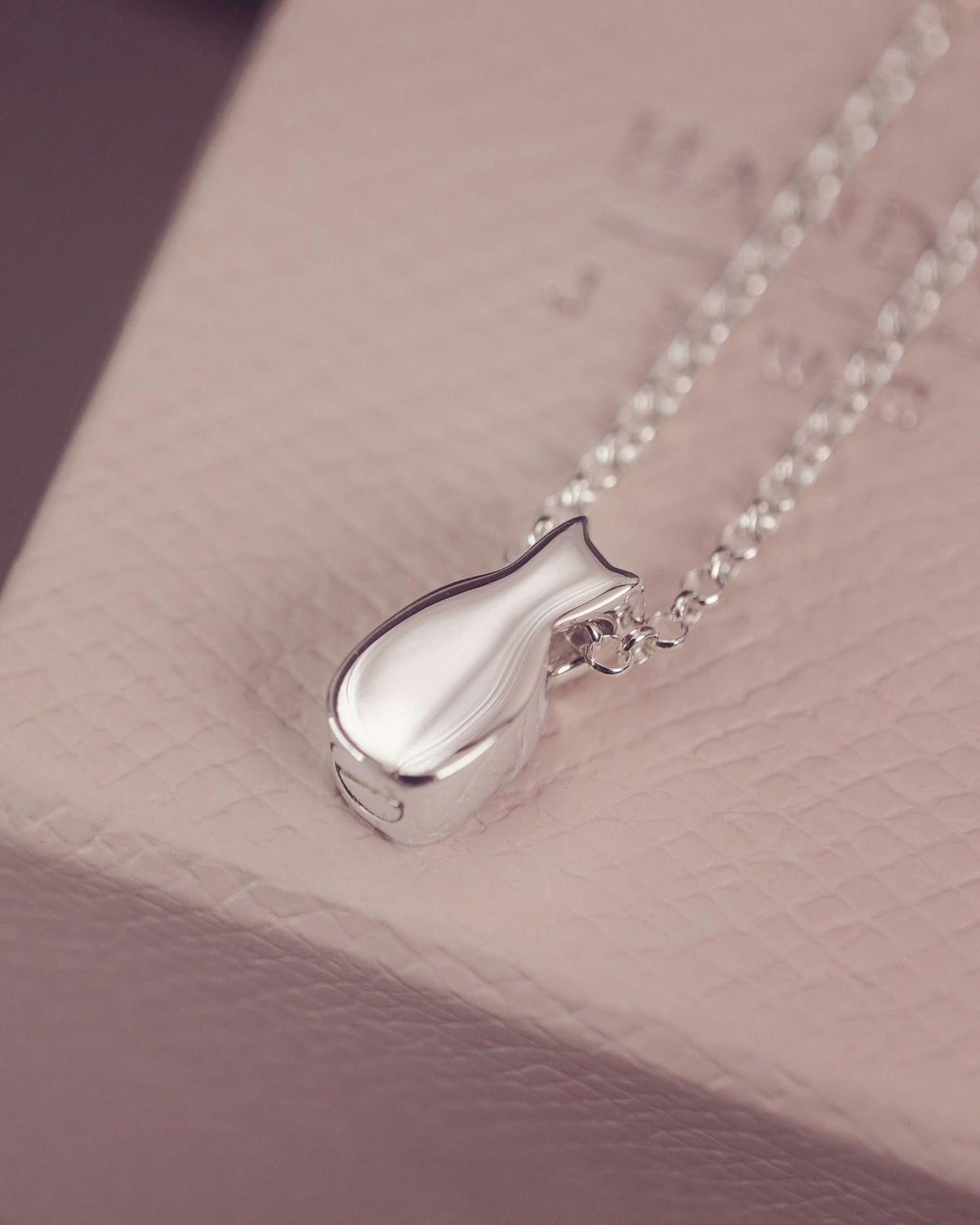 A silver pendant necklace with a cat-shaped charm resting on a textured light pink jewelry box.
