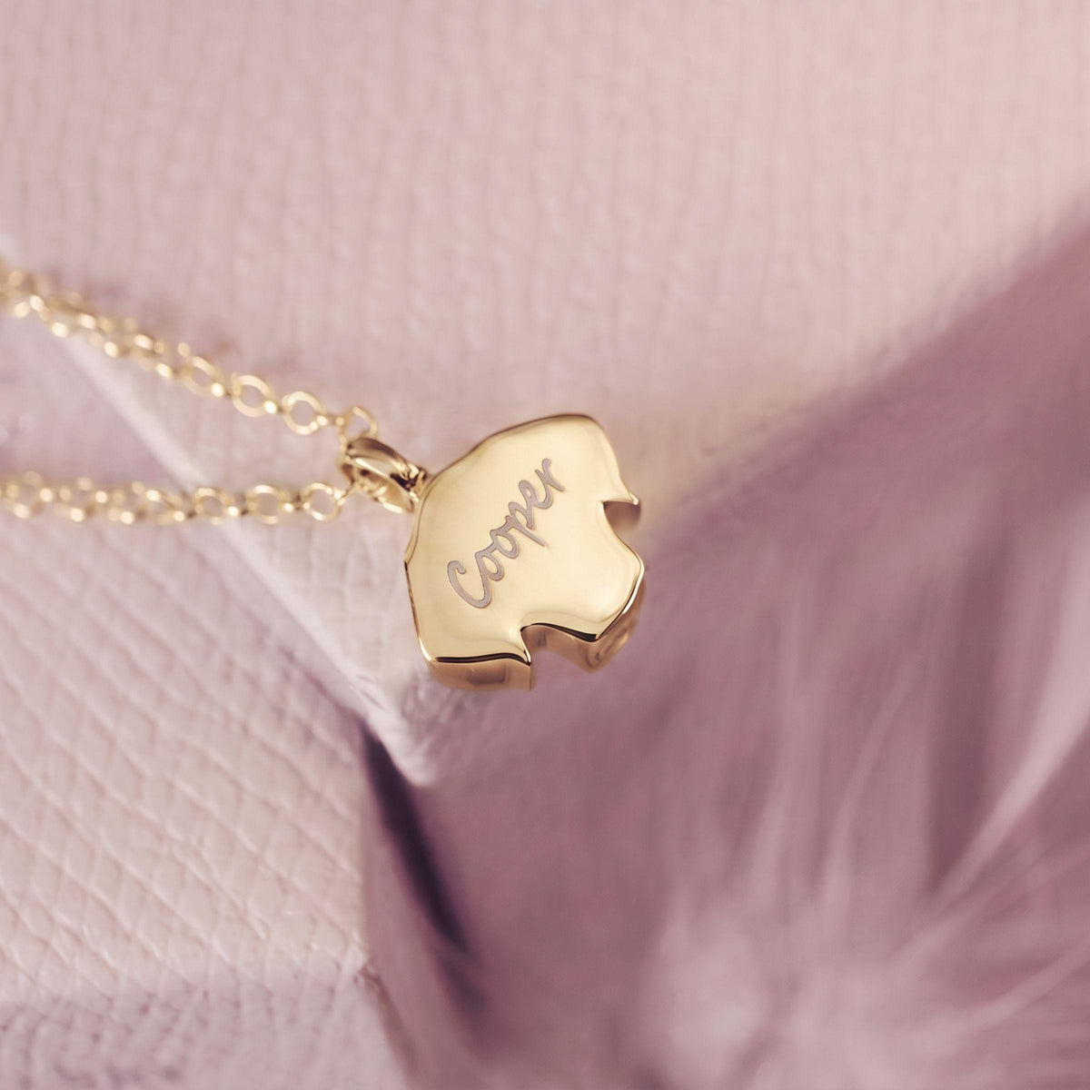 A gold necklace with a dog-shaped pendant engraved with the name 'Cooper', resting on a textured pink surface with a soft feather in the background.