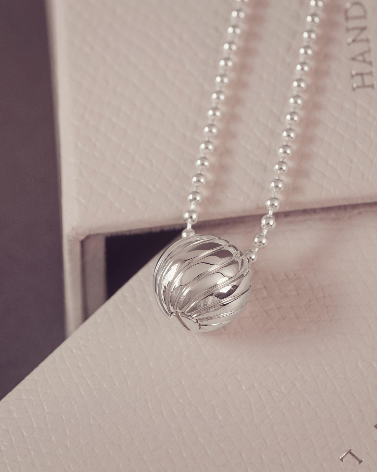 A silver beaded necklace with a textured, ribbed spherical pendant, resting on a light beige textured box.