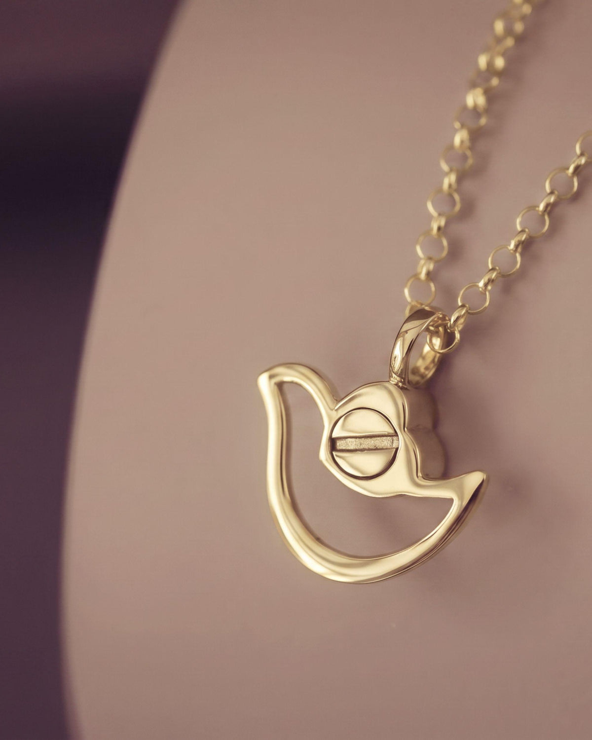 A gold pendant necklace featuring a stylized dove shape with a central screw-like detail, shown resting on a soft beige display surface against a dark purple background.