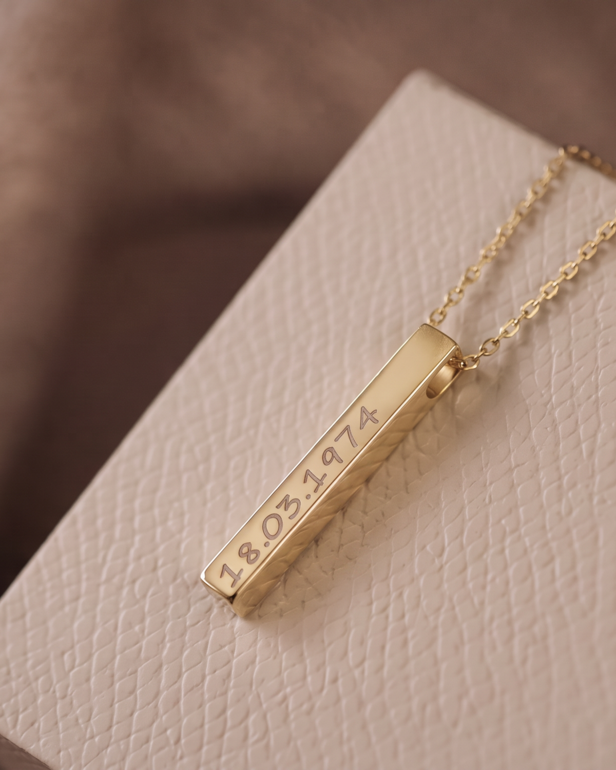 A gold bar pendant necklace engraved with the date '18.05.1974', resting on a textured cream-colored gift box.