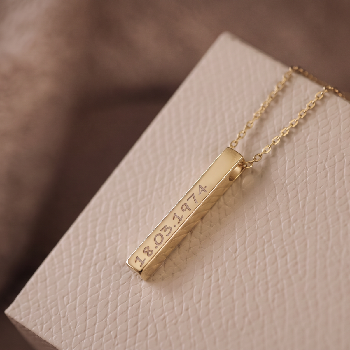 A gold bar pendant necklace engraved with the date '18.05.1974', resting on a textured cream-colored gift box.