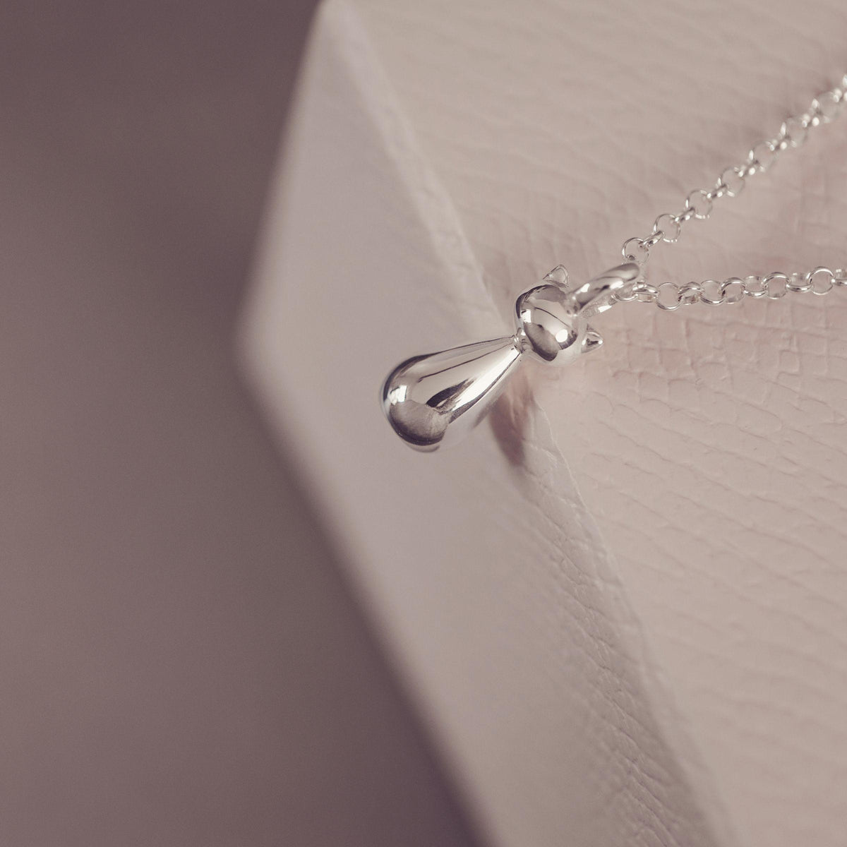 A close-up of a silver stylised cat pendant necklace on a delicate chain, resting against a textured white display block.
