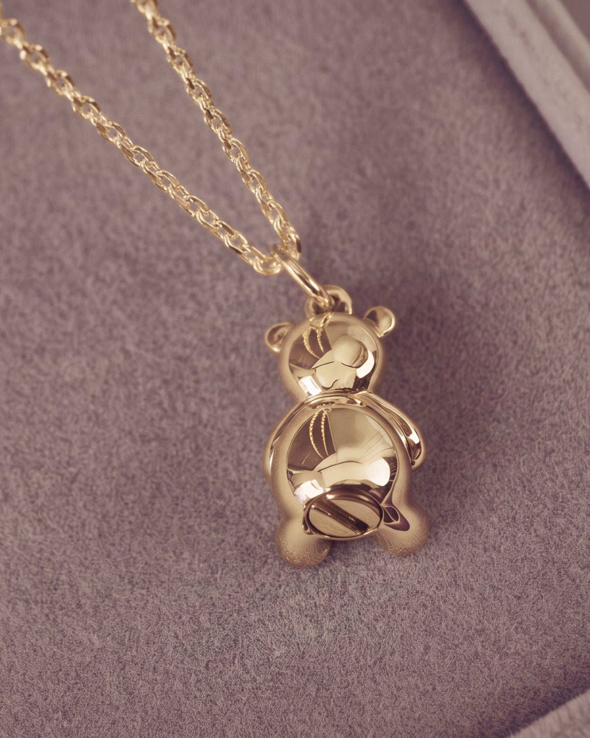 A gold teddy bear pendant necklace resting on a textured light jewellery box, showing the back of the bear and a portion of the chain.