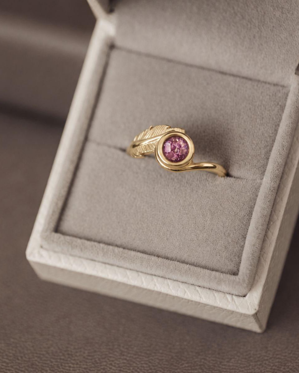 A gold ring with a round pink gemstone and a leaf-like textured accent on the band, displayed inside an open light gray jewelry box.