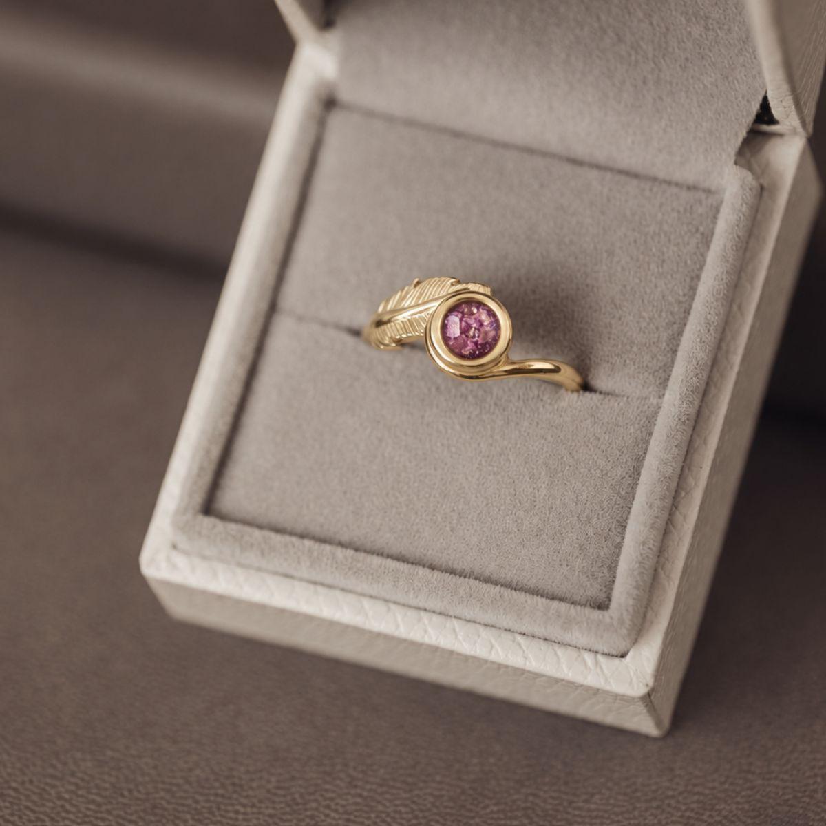 A gold ring with a round pink gemstone and a leaf-like textured accent on the band, displayed inside an open light gray jewelry box.