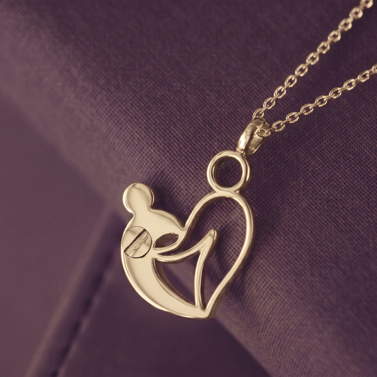 A gold pendant necklace featuring a stylized mother-and-child silhouette design, resting on a textured purple fabric background.