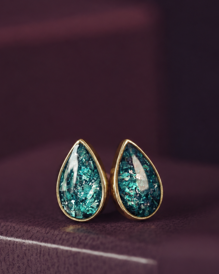 A pair of gold teardrop stud earrings with textured, glittery teal-green resin centers, resting on a dark purple surface.