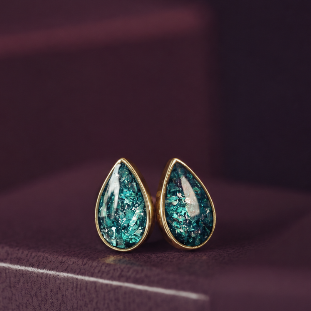 A pair of gold teardrop stud earrings with textured, glittery teal-green resin centers, resting on a dark purple surface.