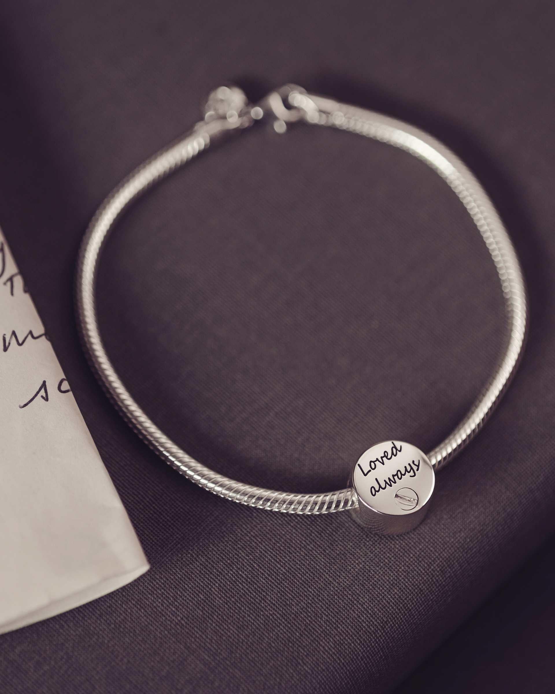 Round bracelet urn charm engraved with 'loved always' sitting on a thick snake chain bracelet.dark background along side a letter.