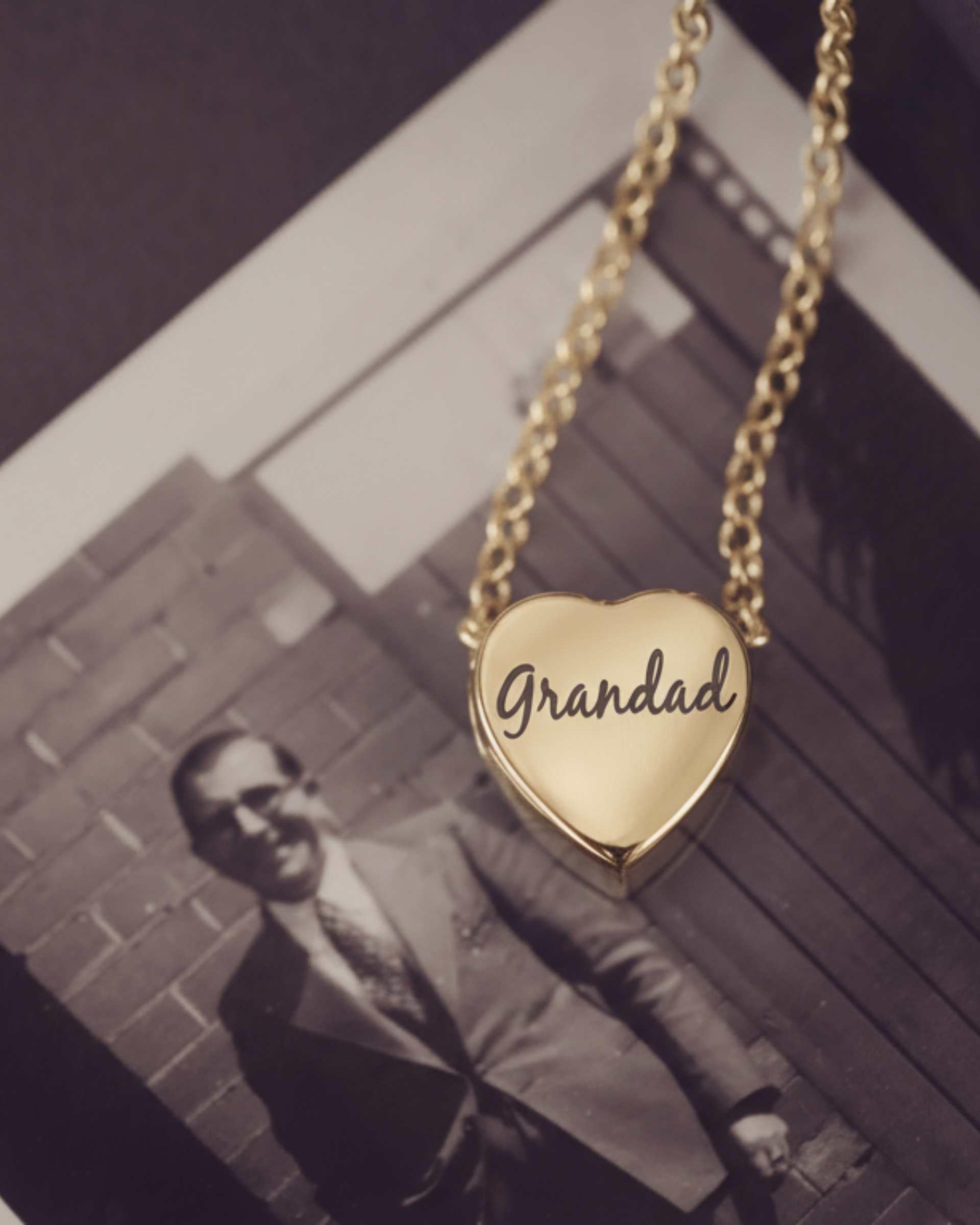 Gold heart-shaped pendant with 'Grandad' engraved, hanging above a black and white photo of a man.