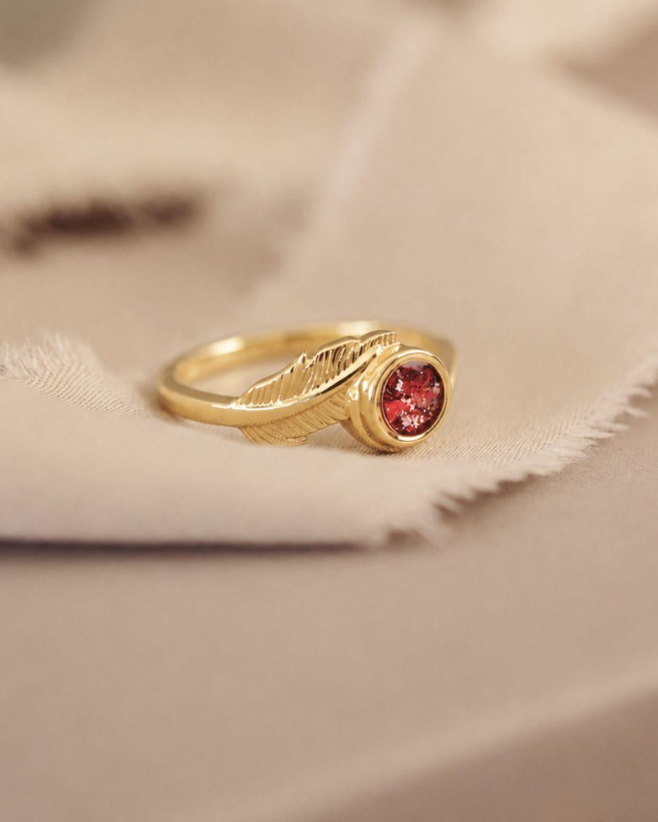A gold ring with a textured leaf-like band and a round red gemstone, resting on a soft beige fabric background.