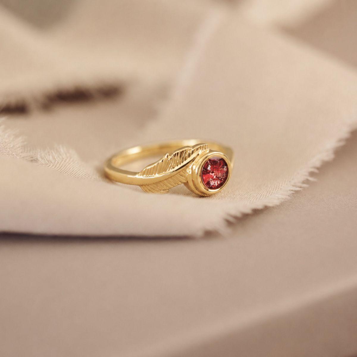 A gold ring with a textured leaf-like band and a round red gemstone, resting on a soft beige fabric background.