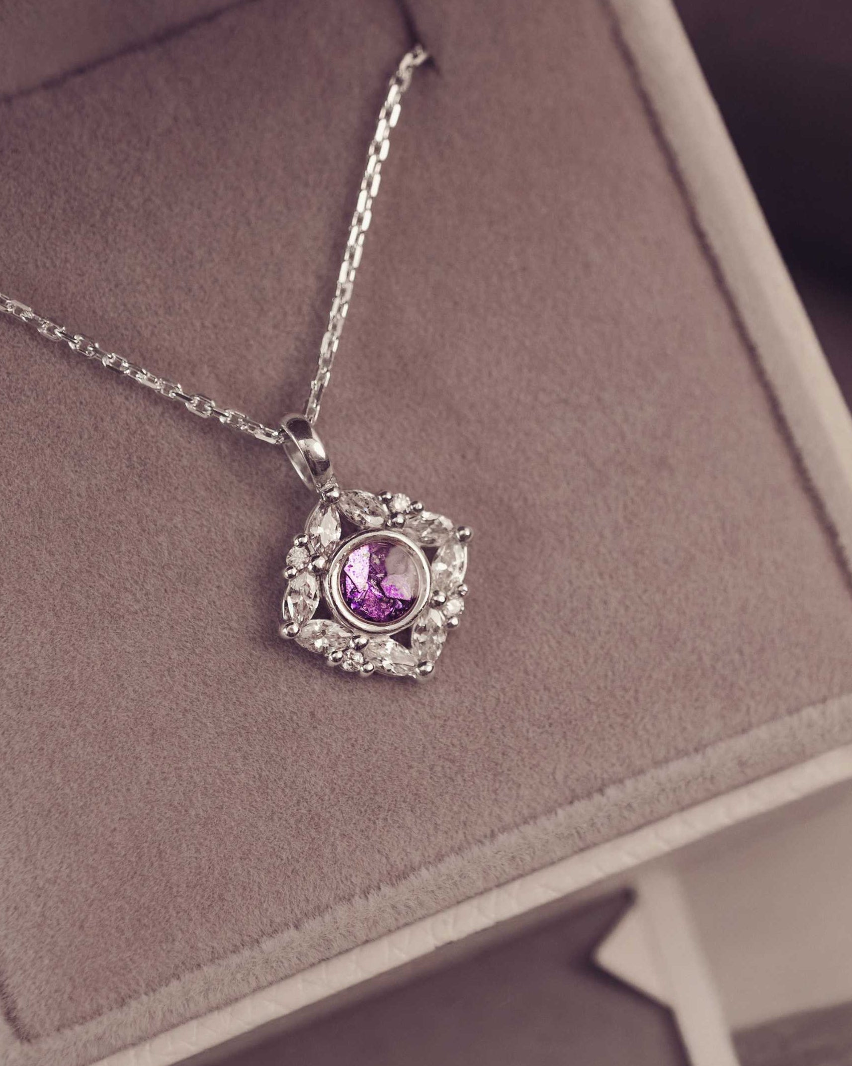 Necklace with a purple gemstone pendant on a pink cushion, with a blurred background of people and nature.