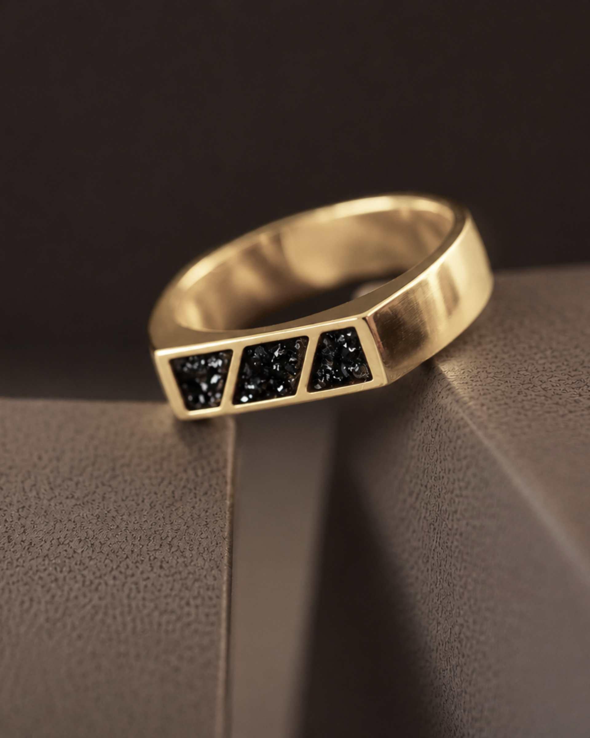 A gold ring with 3 sections of blacked resin across the top. Sitting on a textured dark background