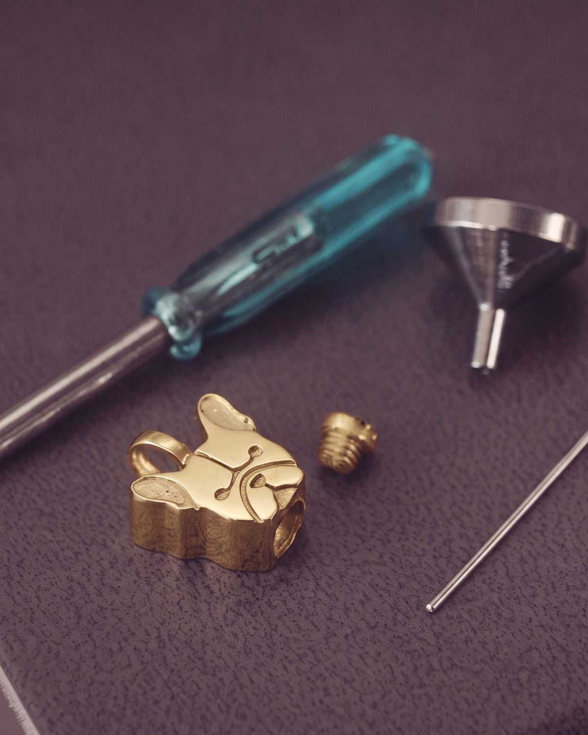 Gold french bulldog pendant charm and small hardware pieces arranged on a textured dark purple surface with a blue-handled screwdriver, funnel, and pin.