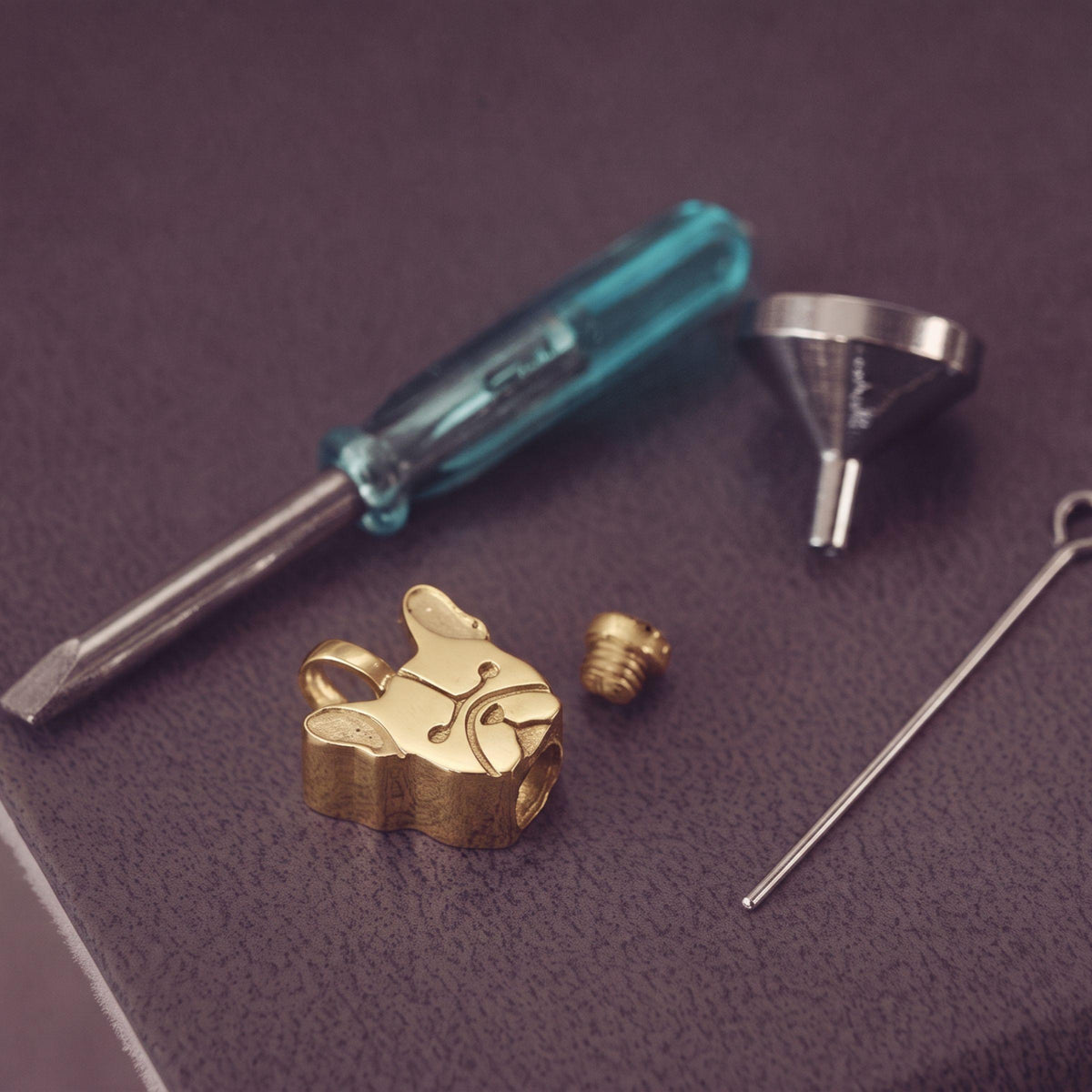Gold french bulldog pendant charm and small hardware pieces arranged on a textured dark purple surface with a blue-handled screwdriver, funnel, and pin.