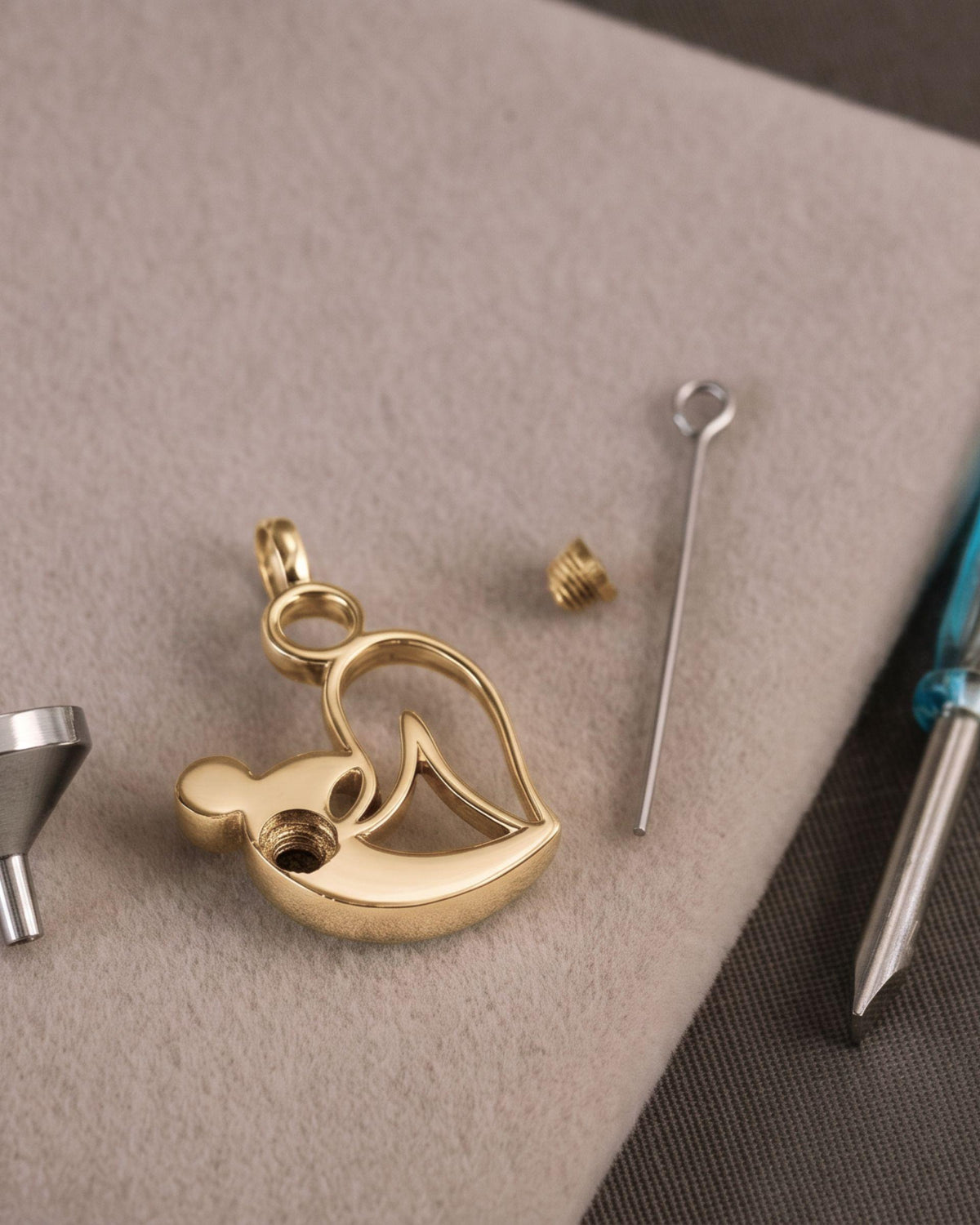 A styalised mother and child pendant charm with a small bail, shown with a silver funnel, a small screw, a pin, and a blue-handled screwdriver on a beige textured surface.