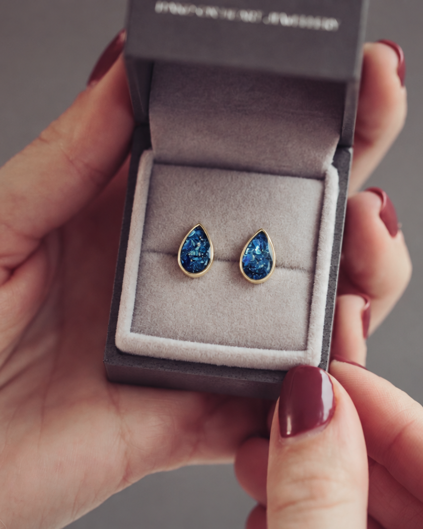 A pair of gold teardrop stud earrings with textured, glittery blue resin centers, resting in a dark grey jewellery box.