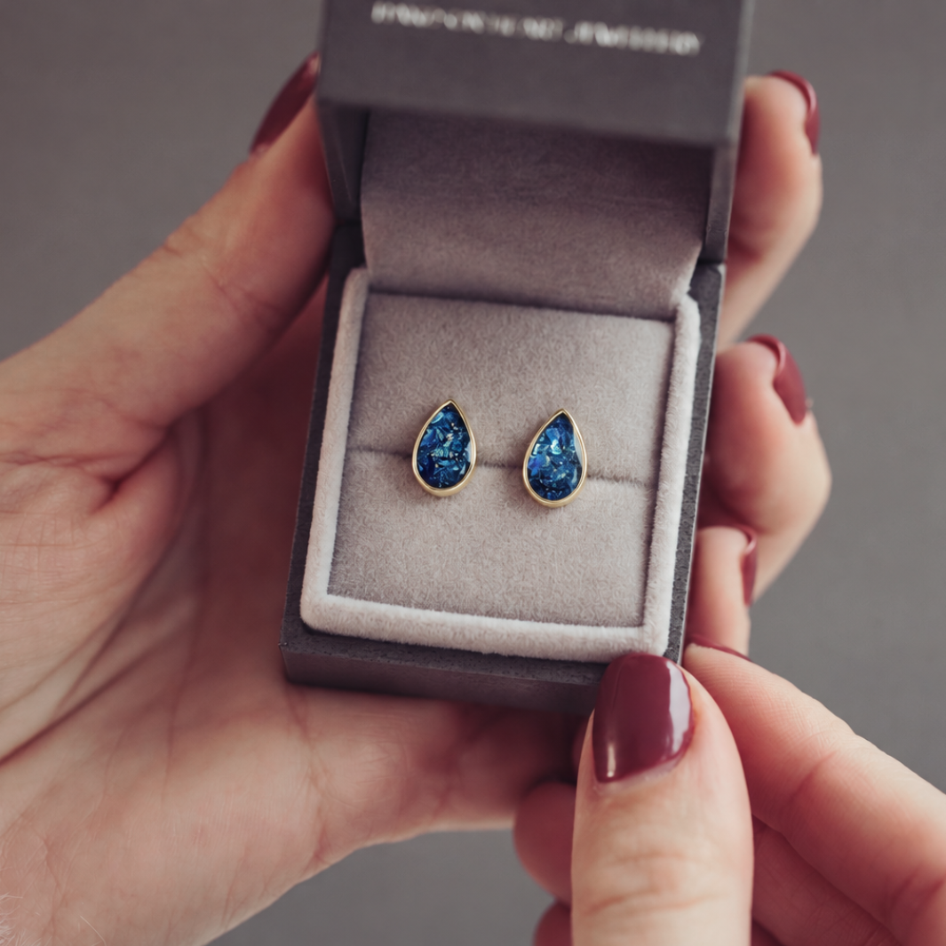 A pair of gold teardrop stud earrings with textured, glittery blue resin centers, resting in a dark grey jewellery box.
