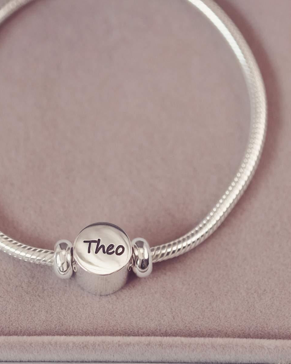 Silver bracelet with a round charm engraved with 'Theo' on a pink background
