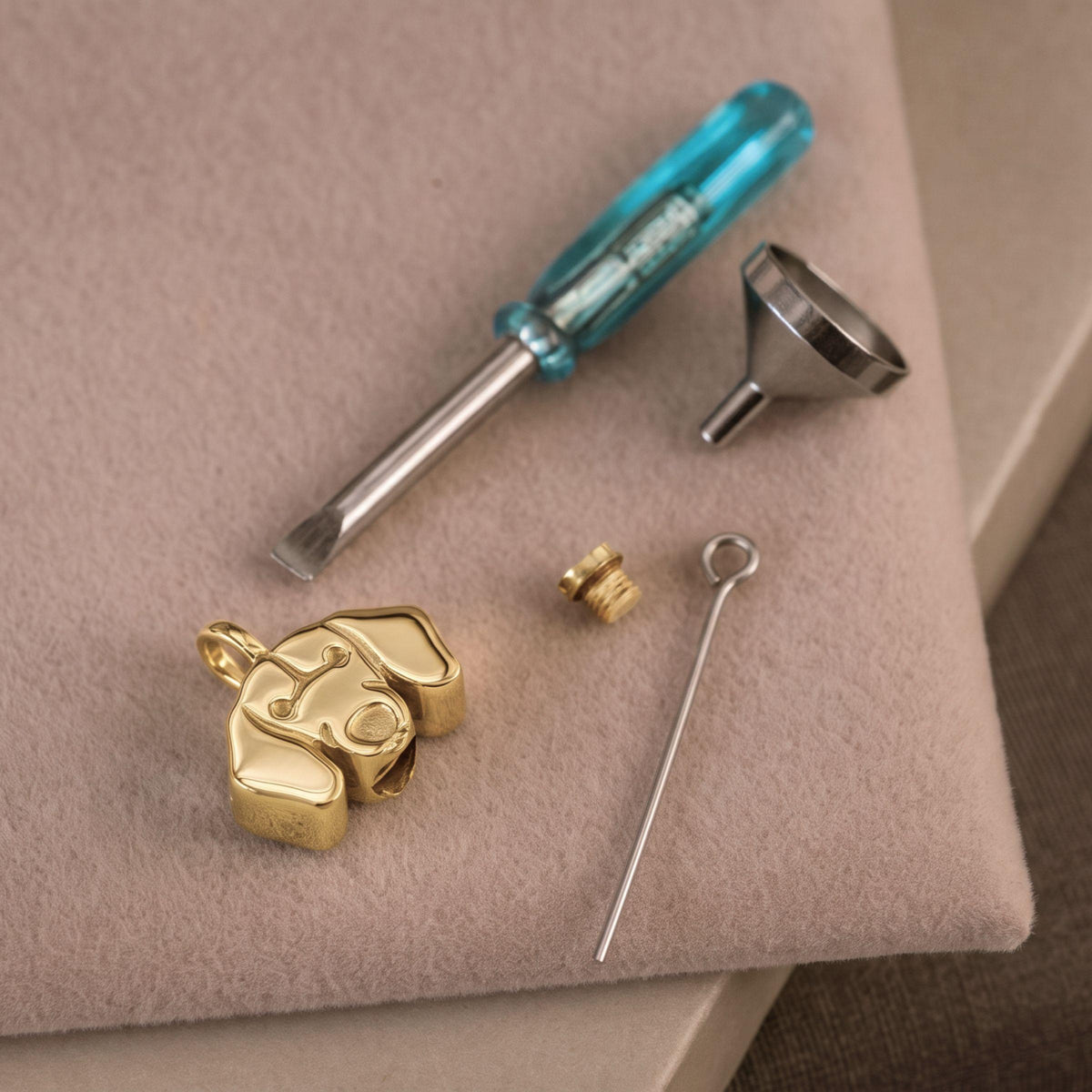 A gold Dachshund dog-shaped pendant charm shown disassembled with a small screwdriver, funnel and pin on a beige felt surface.