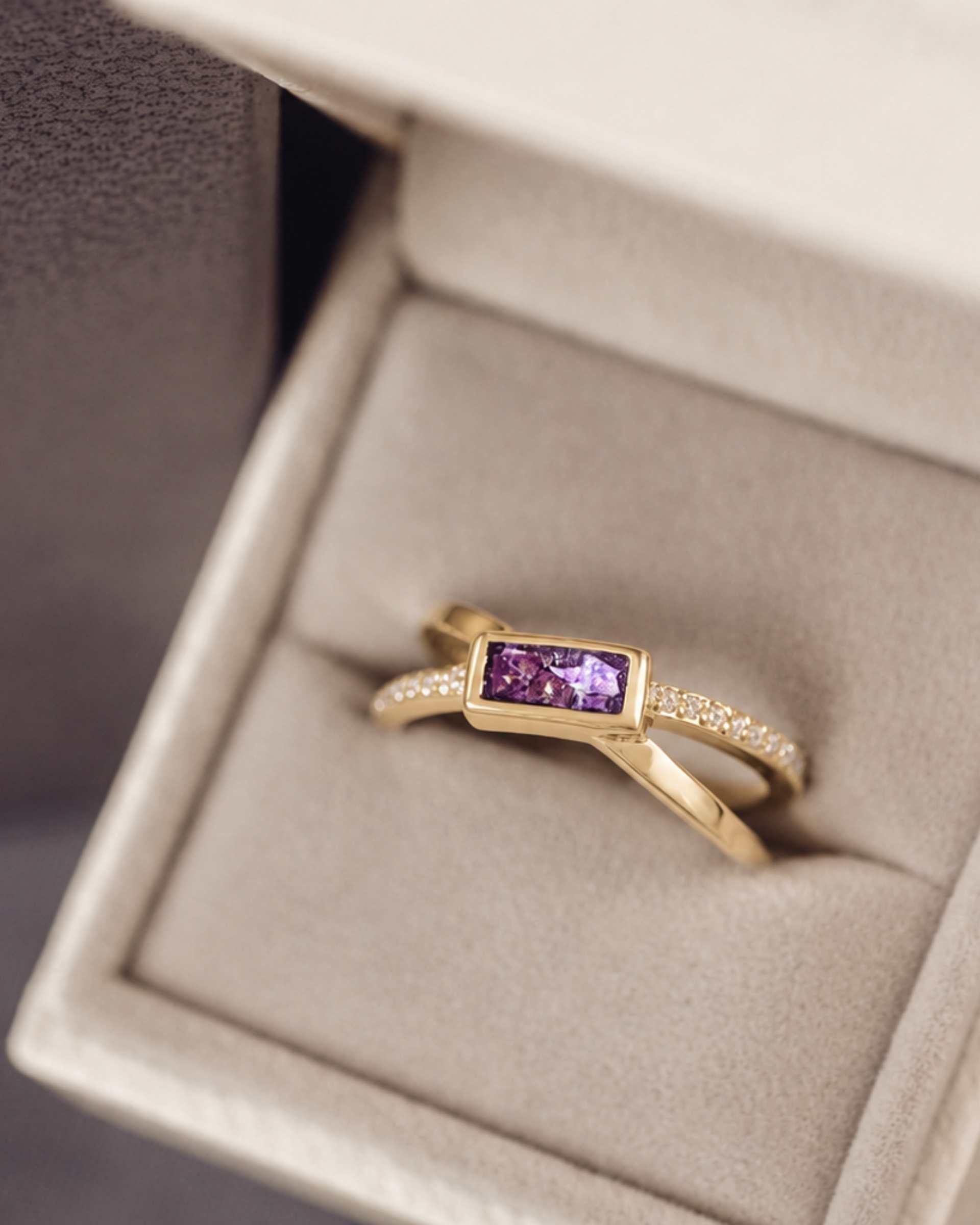 gold criss cross ring with a purple rectangle ash stone and gemstones in a jewellery box