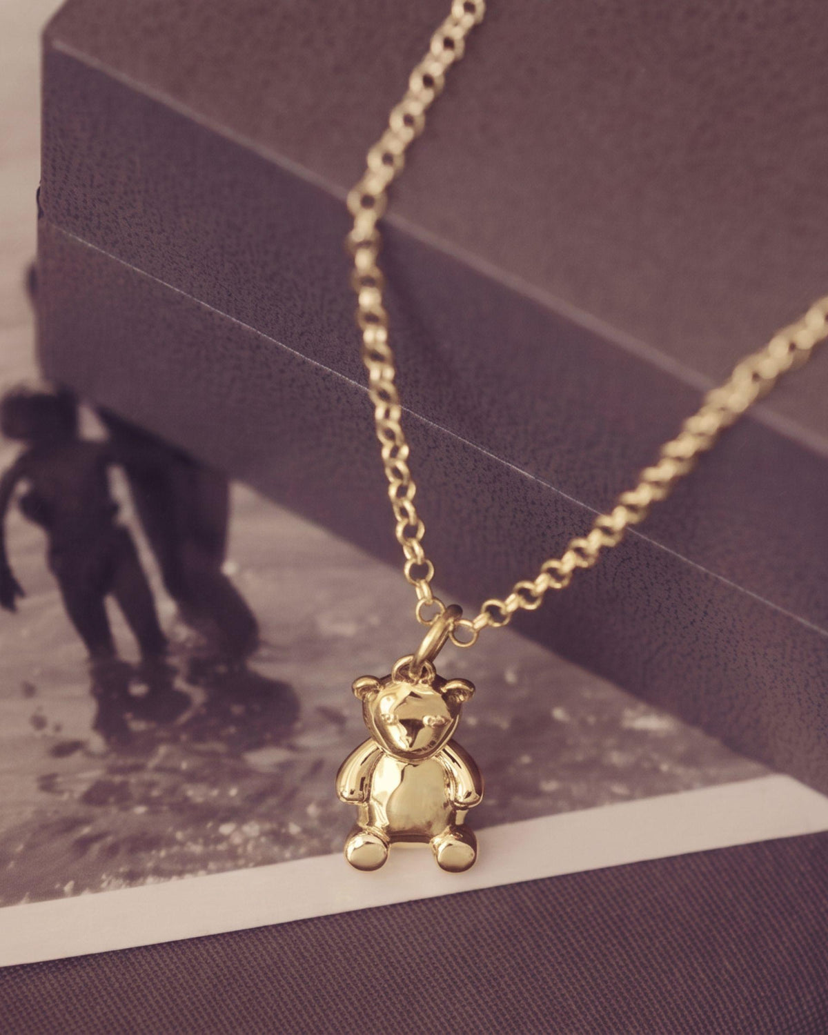 A gold teddy bear pendant necklace on a gold chain, styled on a dark textured box and a vintage black-and-white beach photo print.