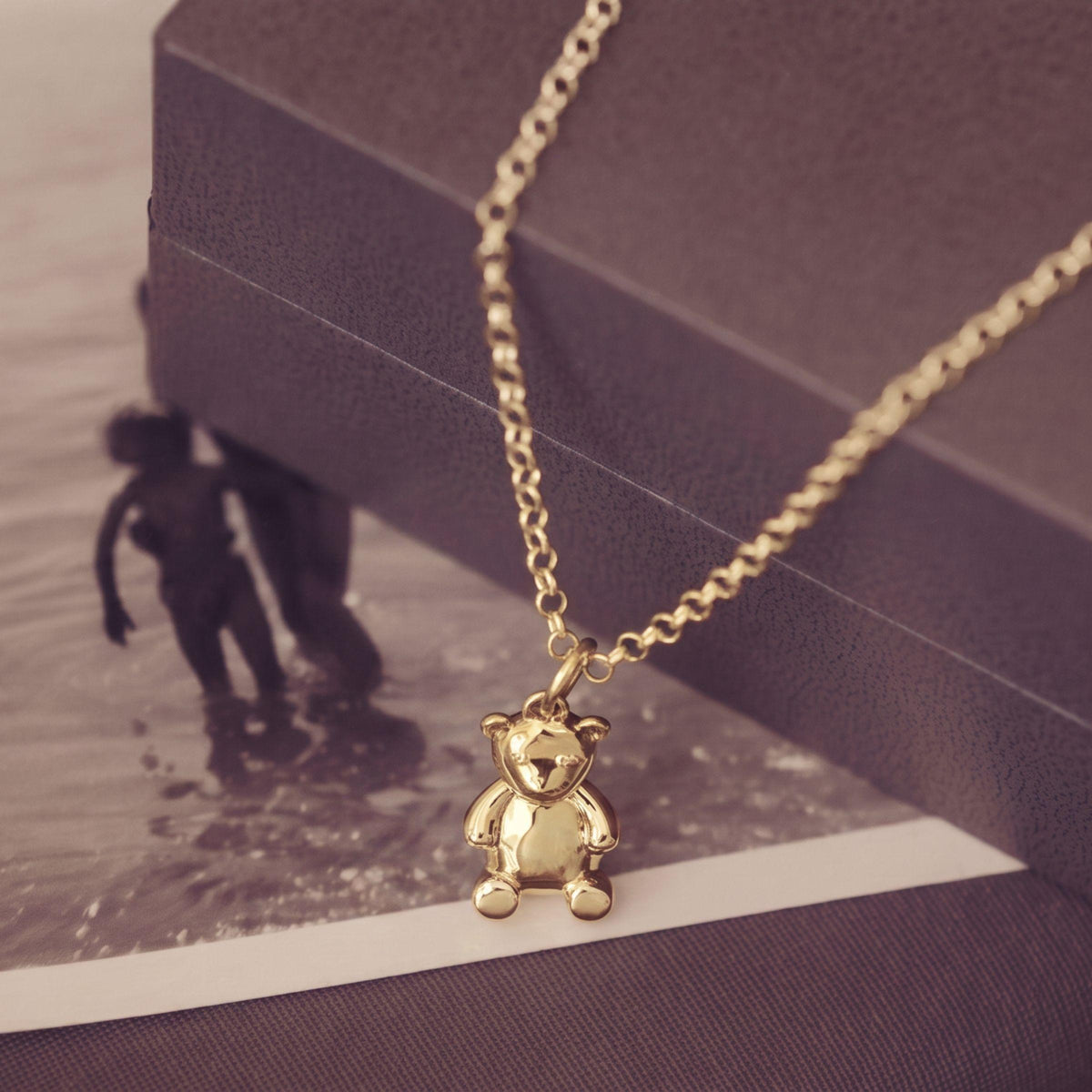 A gold teddy bear pendant necklace on a gold chain, styled on a dark textured box and a vintage black-and-white beach photo print.